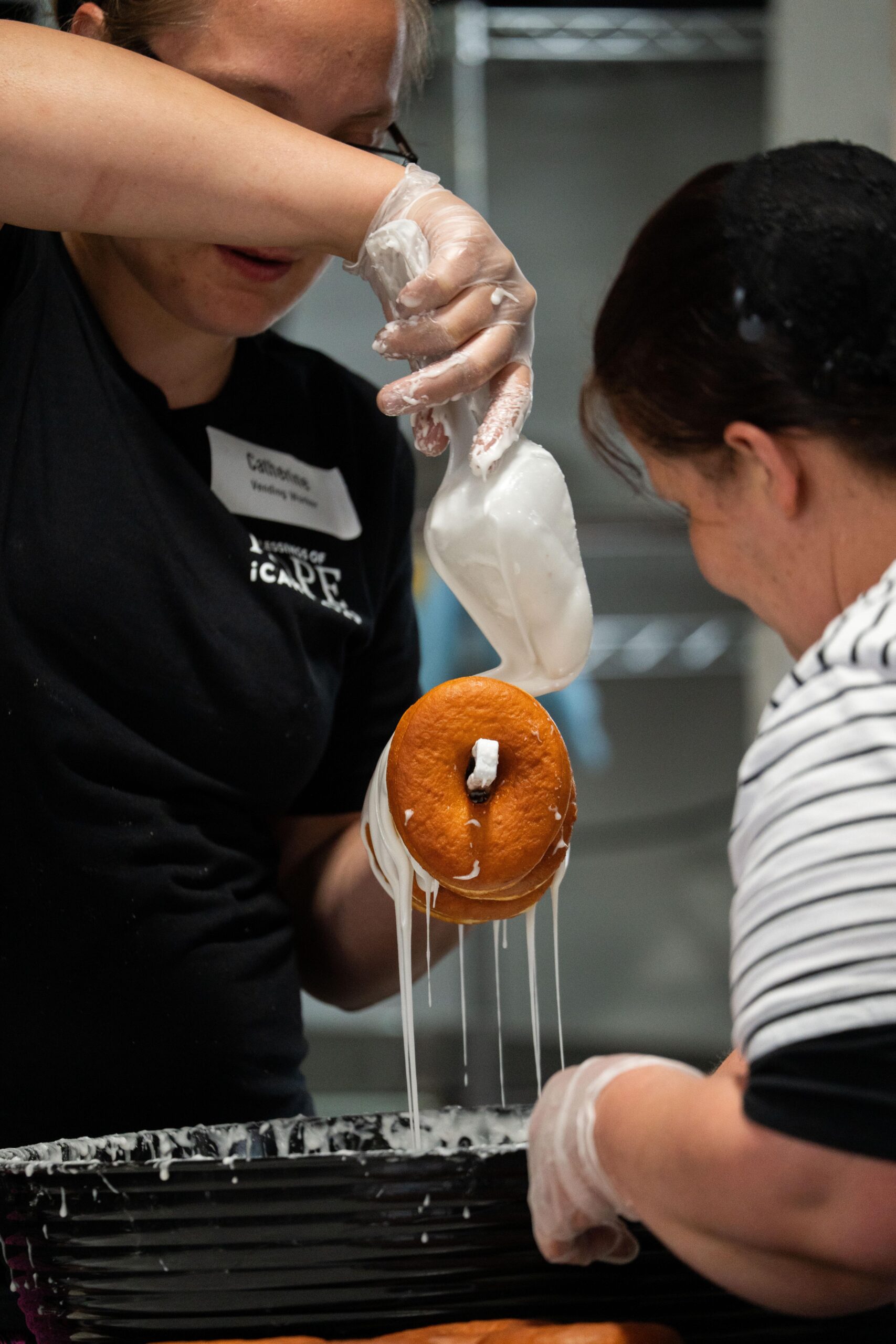 Two individuals are preparing donuts in a kitchen setting, with one person piping icing onto a stacked donut while the other watches. The scene captures a collaborative baking experience with a focus on creativity and culinary skills.