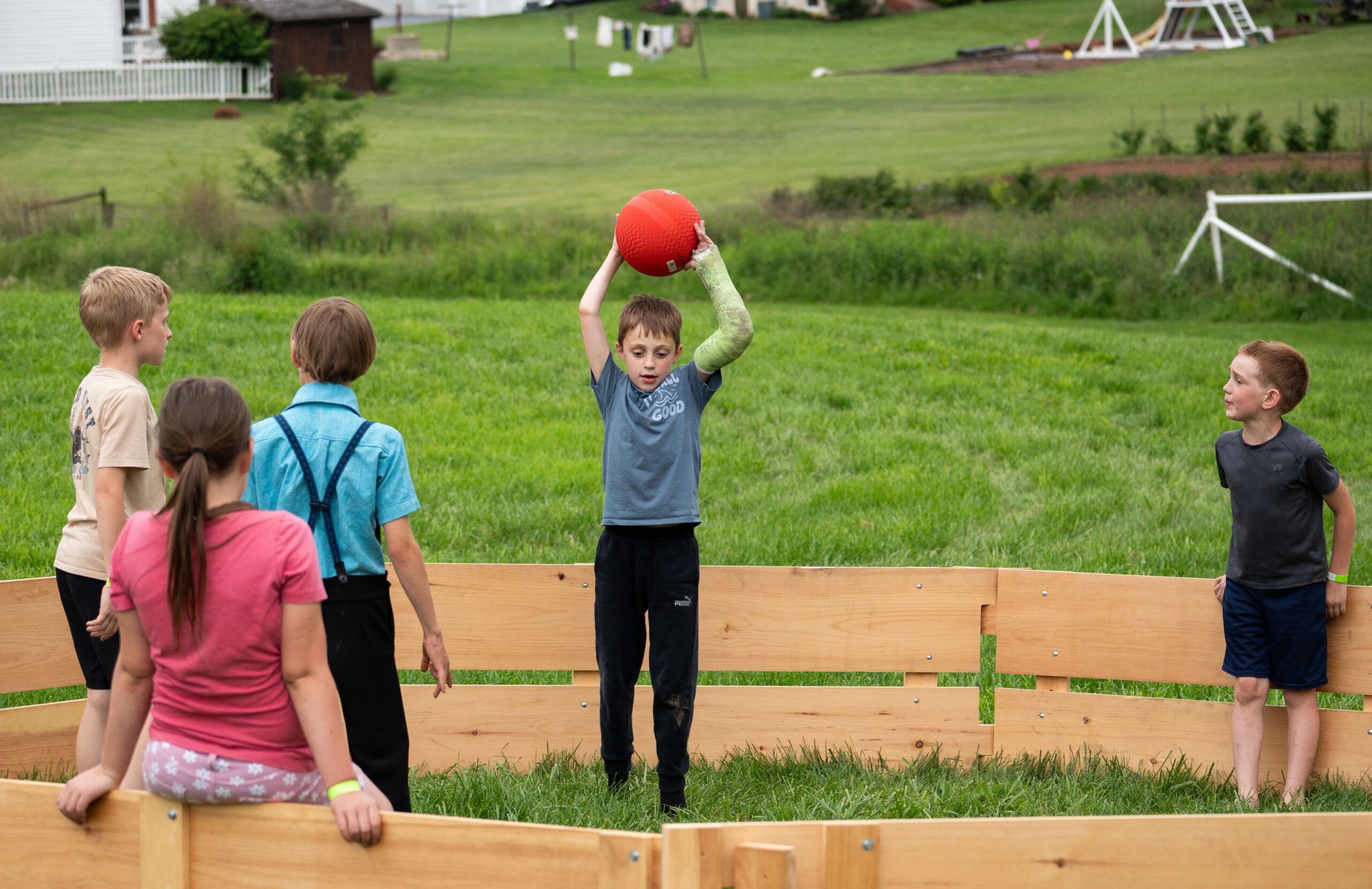 Children playing a game in a wooden fenced area, with one boy wearing a cast holding a red ball above his head while other kids watch. The setting is a grassy yard surrounded by buildings and playground equipment.