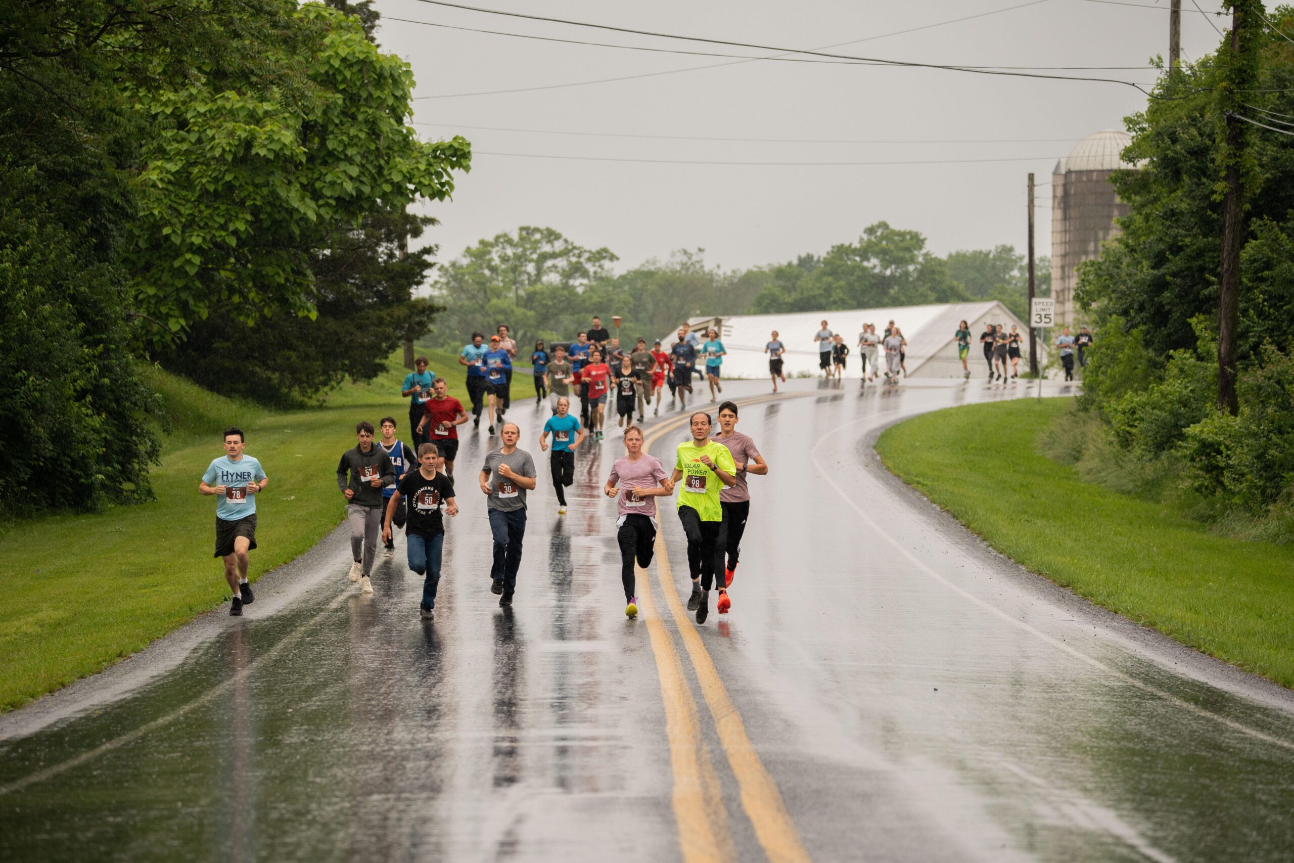 Group of runners participate in a rain-soaked race along a rural road, showcasing determination and athleticism amidst challenging weather conditions. Lush green surroundings and a visible farm structure create a scenic backdrop for the event.