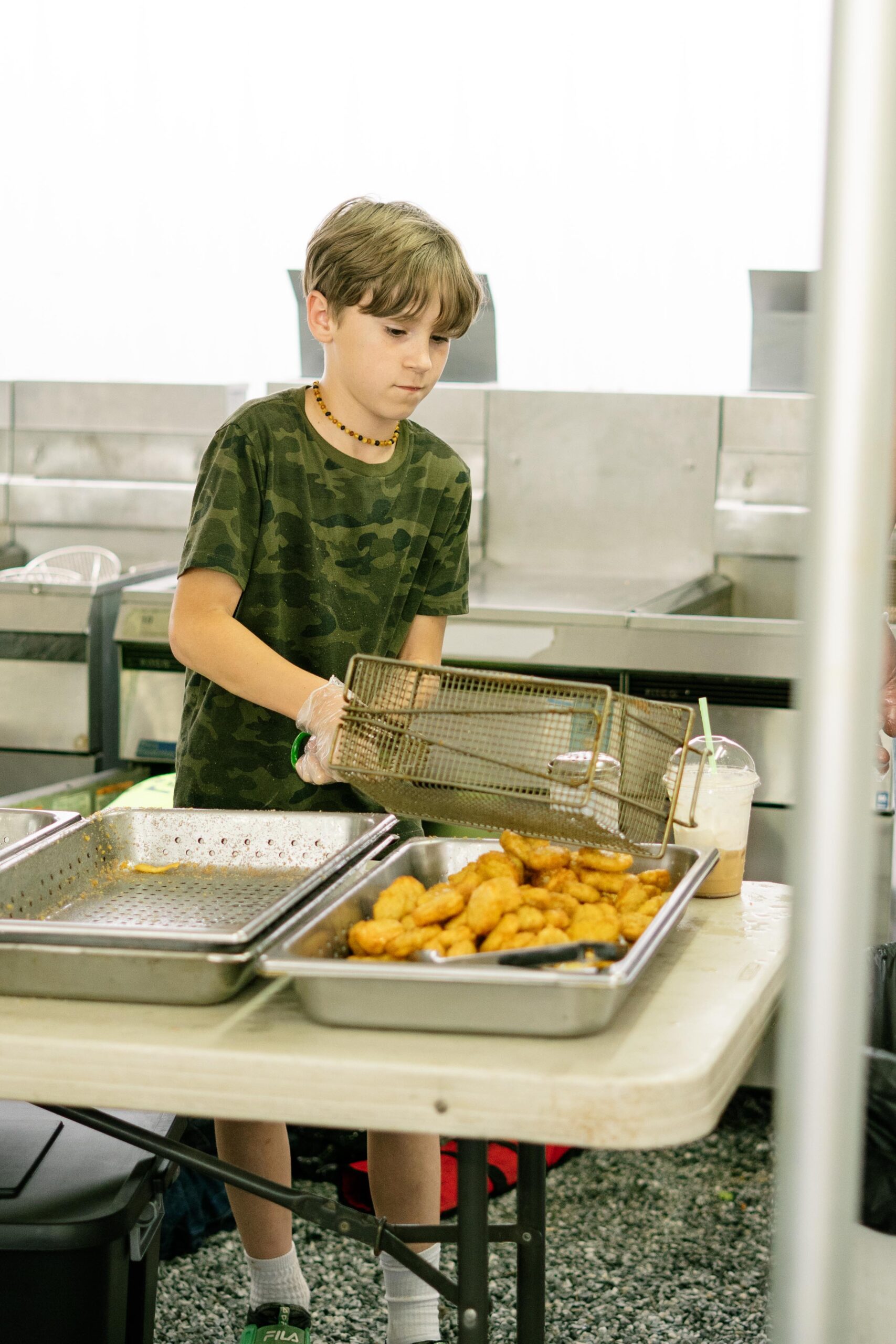 Young boy in camouflage shirt using gloves to serve crispy chicken nuggets from a metal tray in a food preparation area, showcasing a hands-on cooking experience.