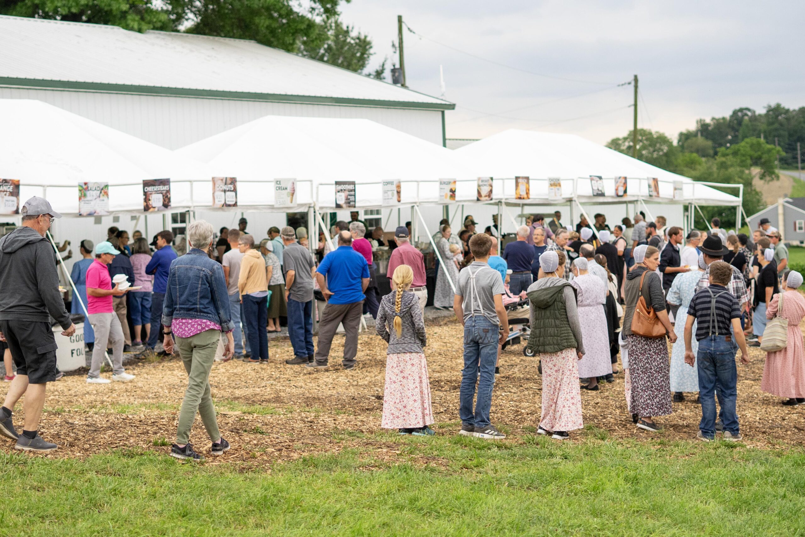 Crowd gathered at a food festival under white tents, enjoying various offerings such as ice cream, donuts, and salads, with attendees of all ages, many dressed in traditional clothing, in a rural outdoor setting.