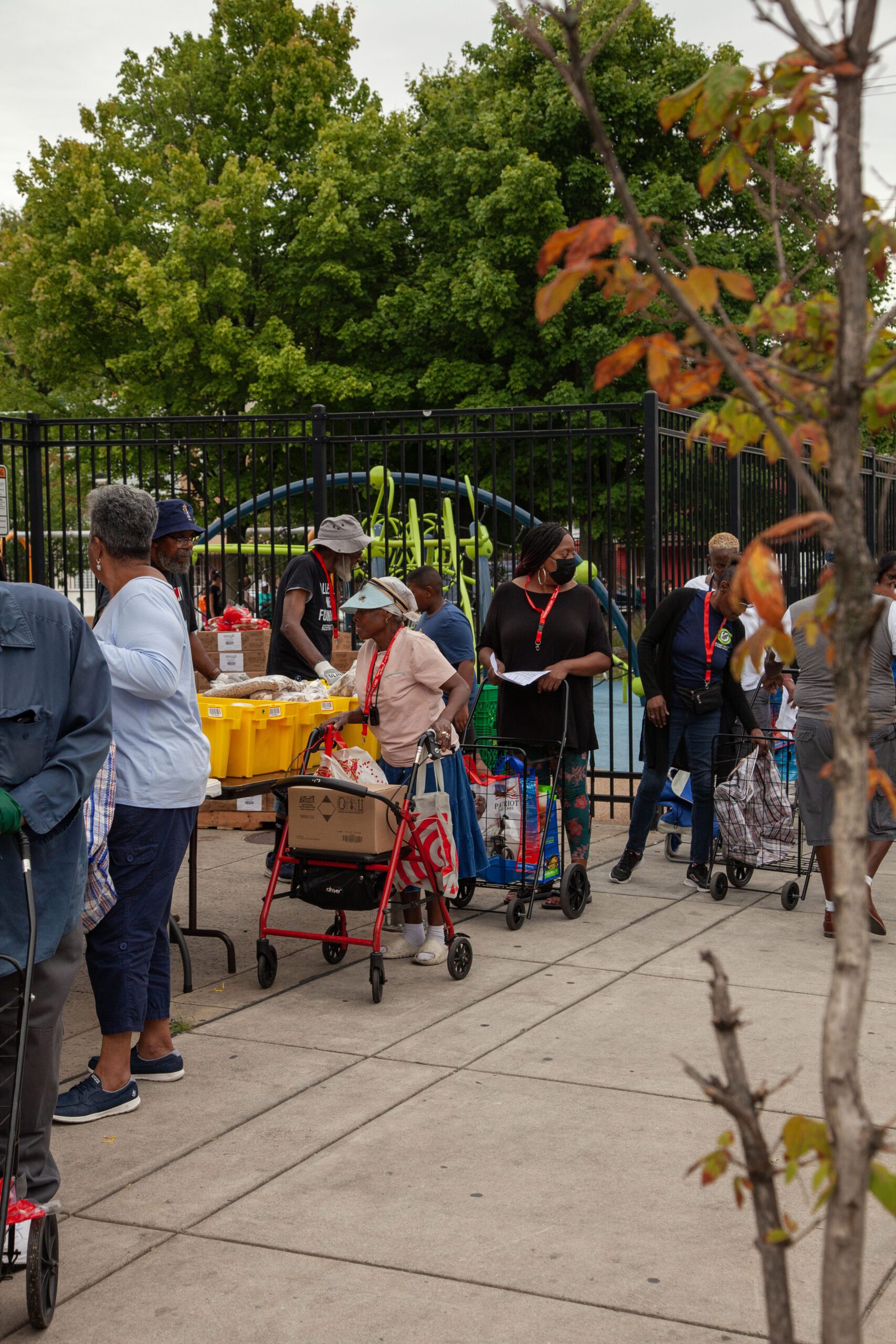 Community members wait in line to receive food assistance, using shopping carts and bags. The scene is set in an outdoor area with trees and playground equipment in the background, reflecting community engagement and support initiatives.