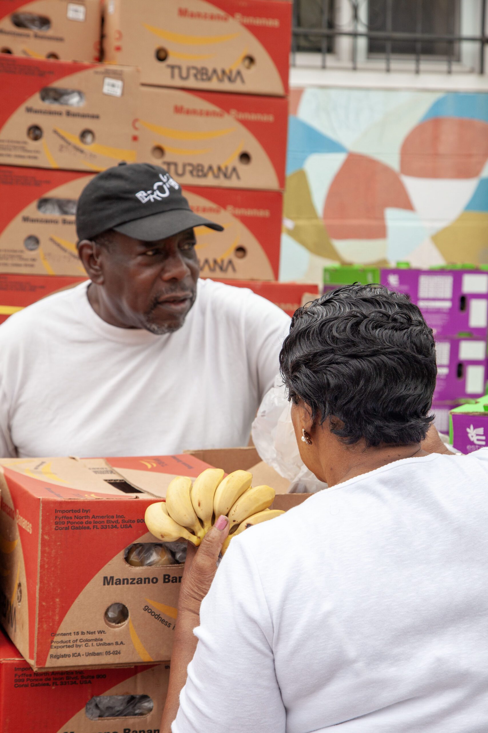 A man in a black hat talks to a woman holding bananas in front of colorful boxes labeled "Turbana Manzano Bananas," showcasing a community market scene focused on fresh produce and local engagement.