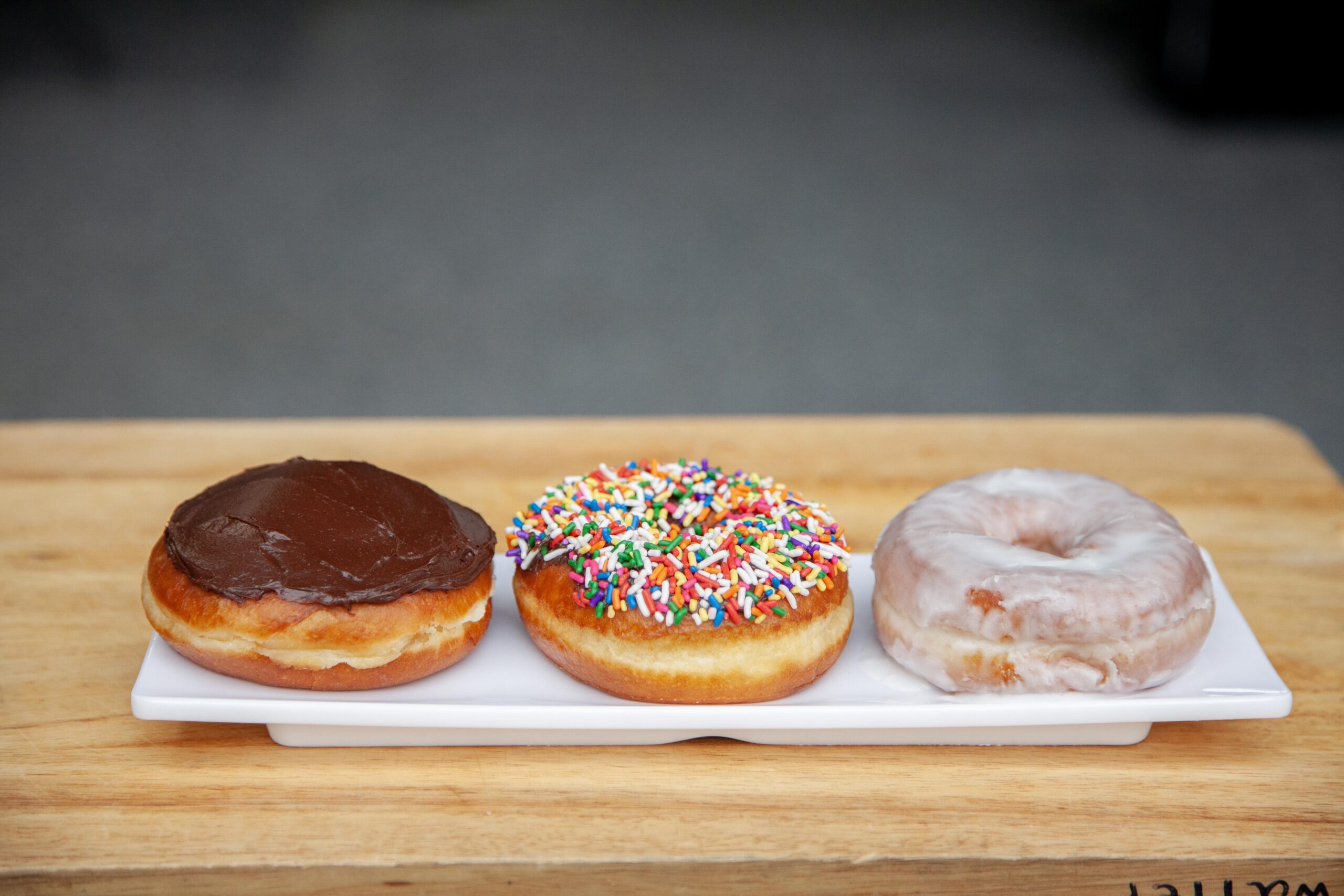 Three assorted donuts on a white platter: a chocolate frosted donut, a colorful sprinkle donut, and a glazed donut, all displayed on a wooden surface. Perfect for a sweet treat or dessert option.