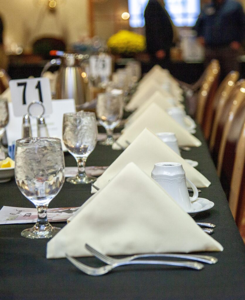 Elegant dining table set for an event, featuring neatly folded napkins, water glasses, and place cards, with a focus on a polished black tablecloth and silver utensils.