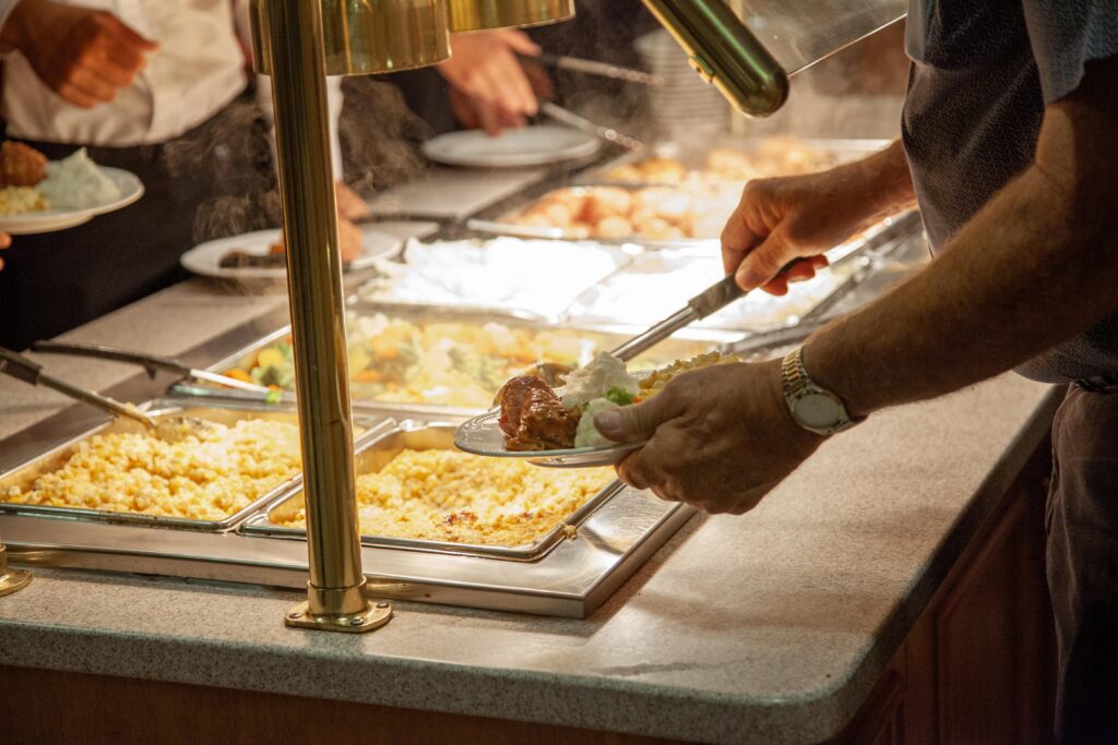 A person serving food from a buffet, selecting a plate of rice and meat while surrounded by various dishes, including vegetables and casseroles, in a warm dining environment.