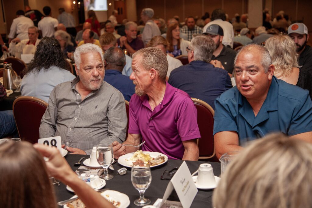 Group of three men engaged in conversation at a dining table during a social event, with plates of food and water glasses in front of them. The setting is lively, with other attendees visible in the background.