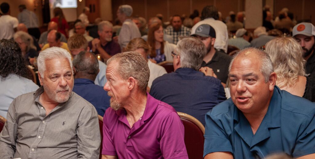 Three men engaged in conversation at a dinner event, with a lively audience in the background. The setting features round tables filled with people, highlighting a social atmosphere.