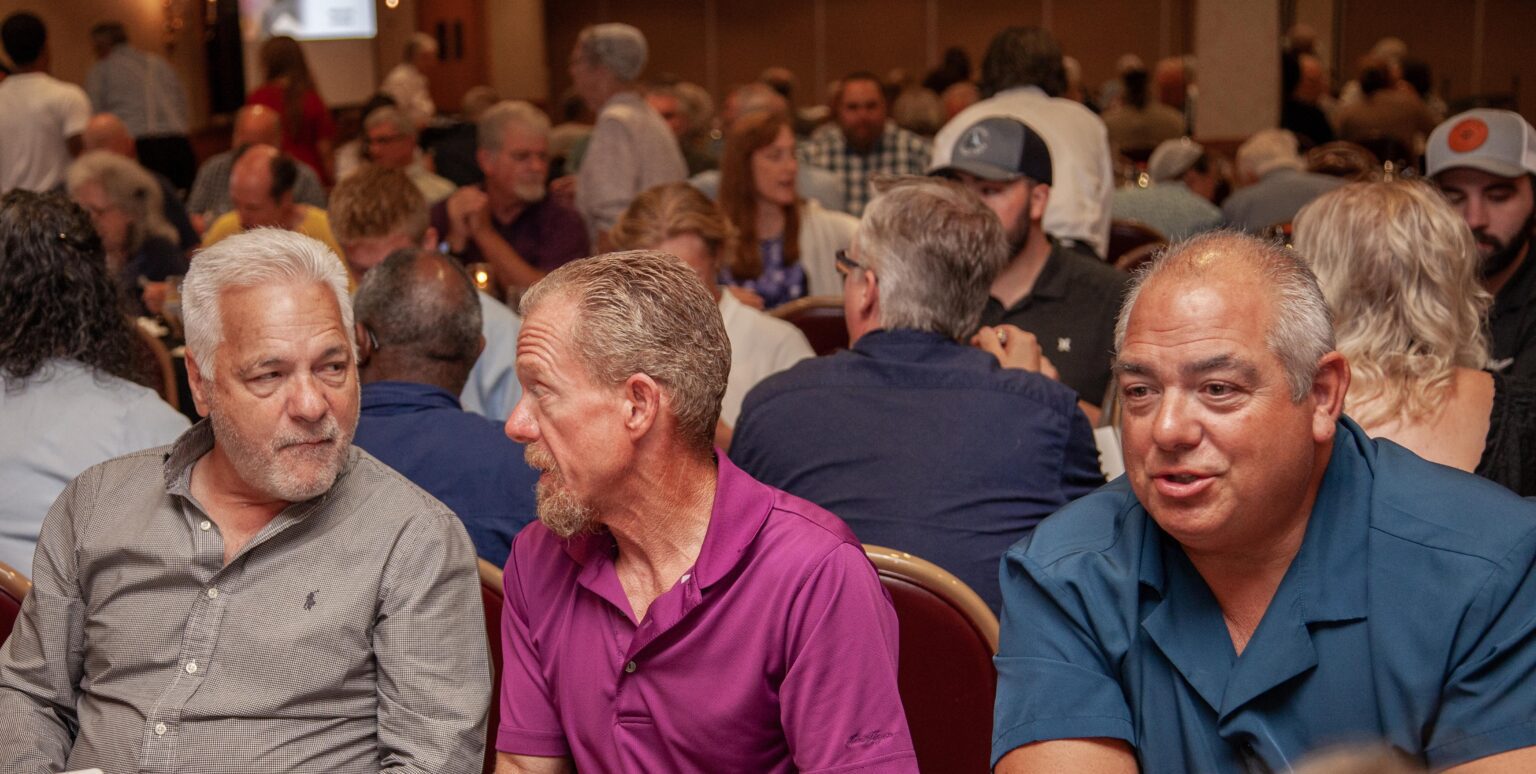 Three men engaged in conversation at a dinner event, with a lively audience in the background. The setting features round tables filled with people, highlighting a social atmosphere.