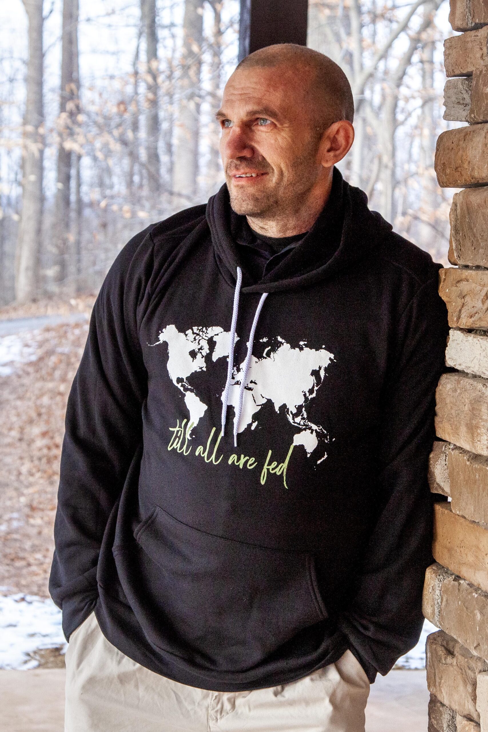 Man wearing a black hoodie with a world map design and the text "till all are fed," standing against a stone wall in a wooded area. The background shows bare trees and a light layer of snow, suggesting a chilly atmosphere.