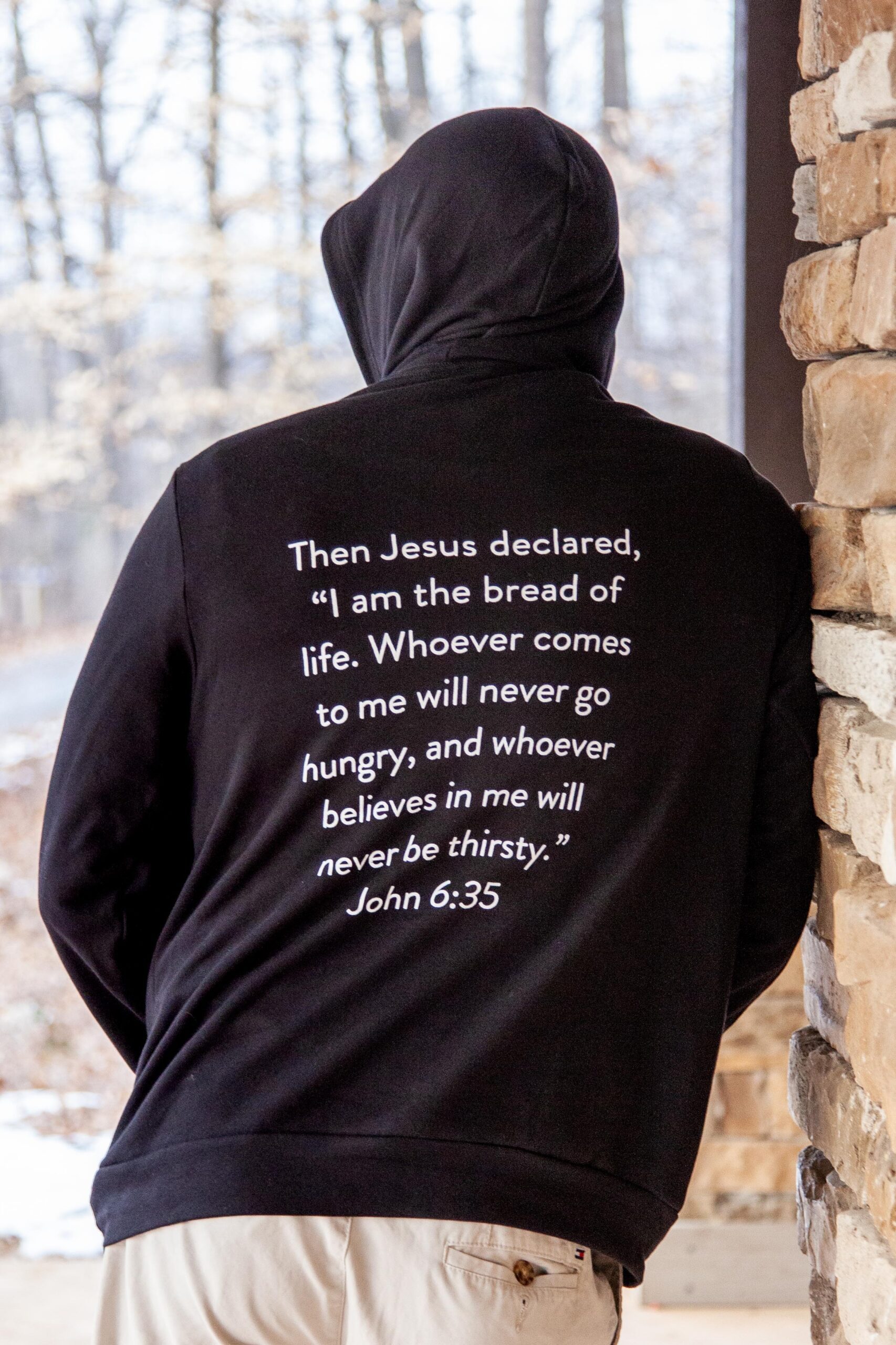 Person wearing a black hoodie with biblical text from John 6:35 printed on the back, standing against a stone wall in a wooded area. The quote emphasizes themes of sustenance and faith.