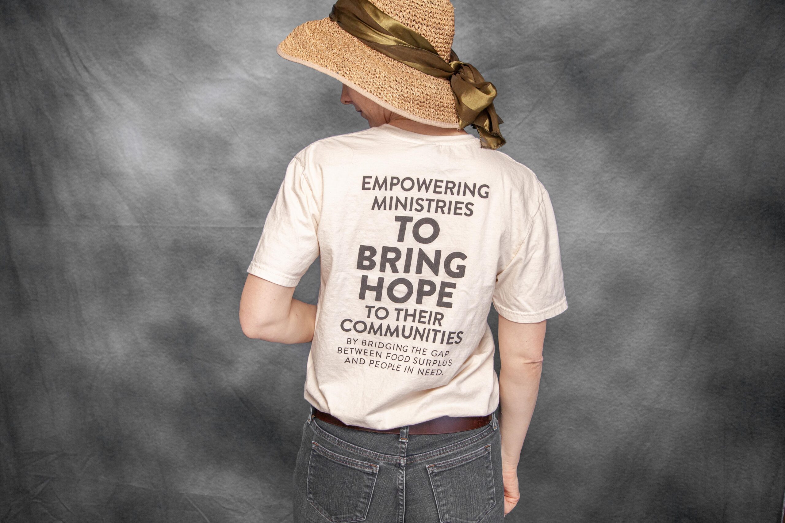 Person wearing a straw hat and a T-shirt with the text "Empowering ministries to bring hope to their communities by bridging the gap between food surplus and people in need," promoting community support and social impact initiatives.