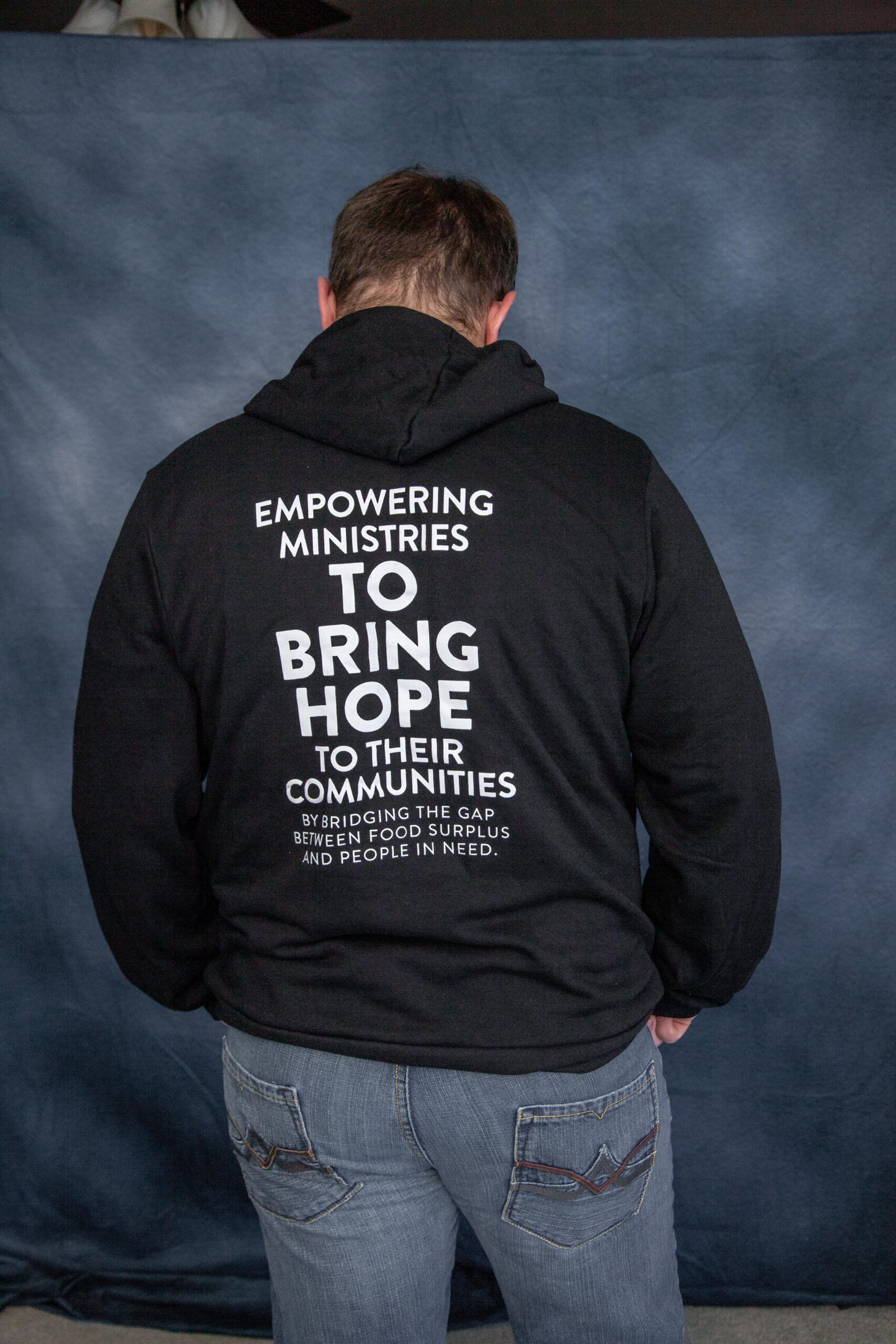 Person wearing a black hoodie with the phrase "Empowering Ministries to Bring Hope to Their Communities" printed on the back, emphasizing food surplus recovery efforts. Background features a soft-focus blue backdrop.