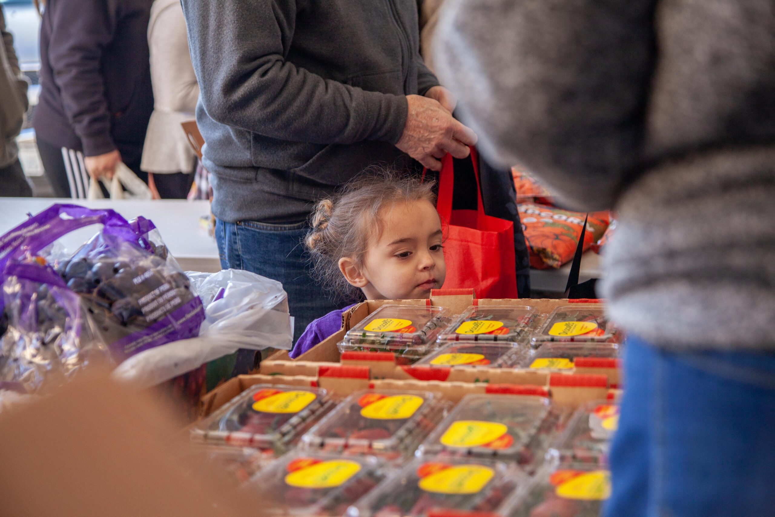A young girl curiously gazes at colorful fruit cartons in a bustling market, surrounded by shoppers carrying bags and examining produce.