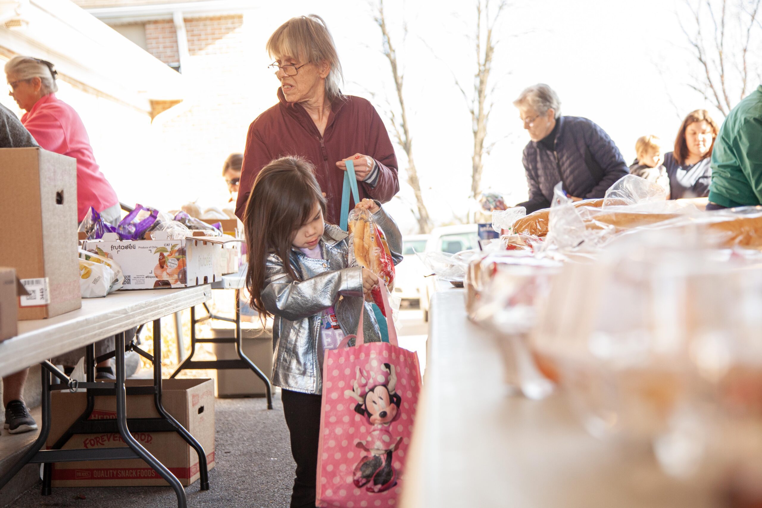 A young girl in a shiny silver jacket is happily picking up items from a table at a community food distribution event, while an elderly woman looks on. Other volunteers and families can be seen participating in the event, promoting community engagement and support.