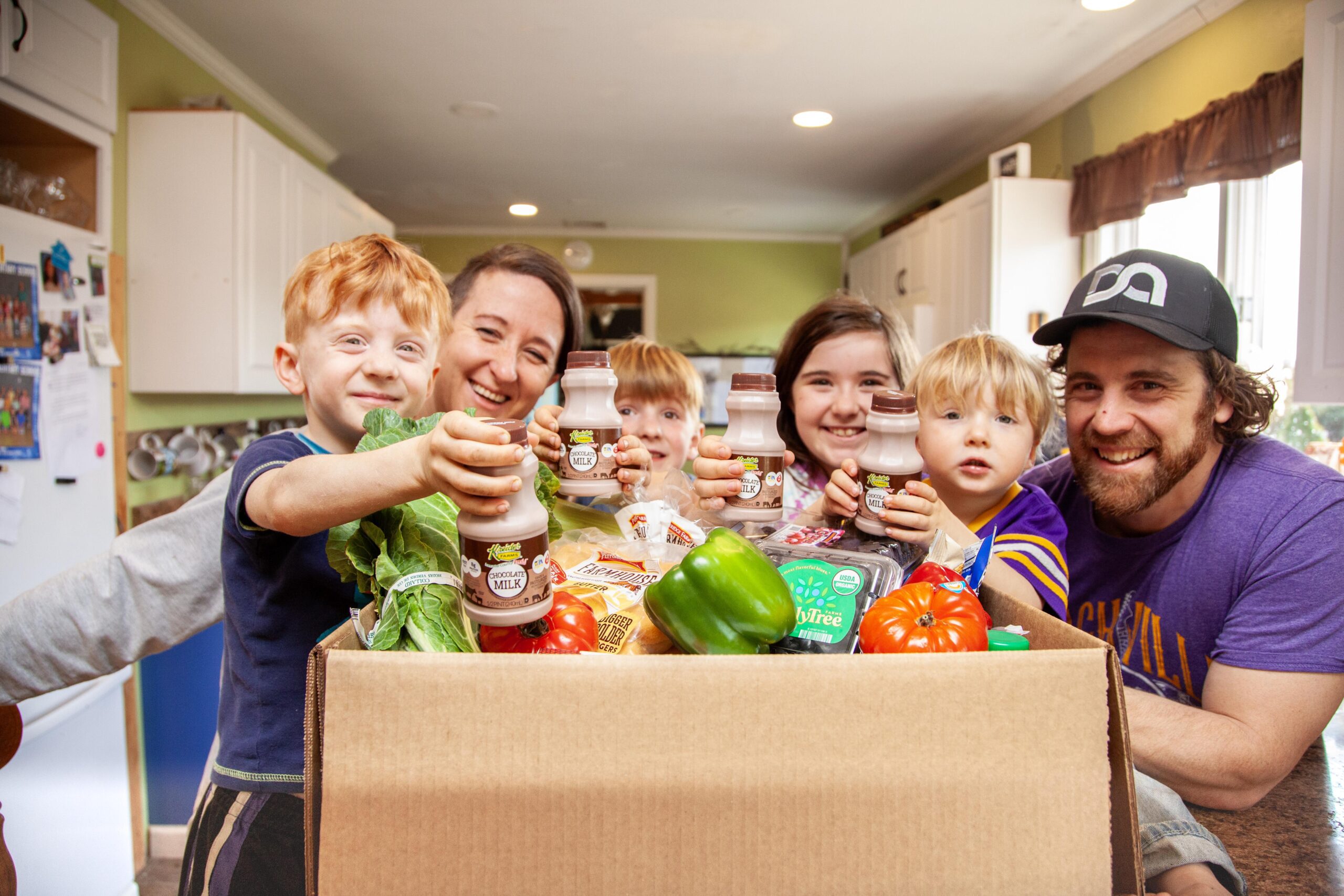 Family joyfully holding healthy groceries, including chocolate milk, fresh vegetables, and fruits, while celebrating healthy eating habits at home.