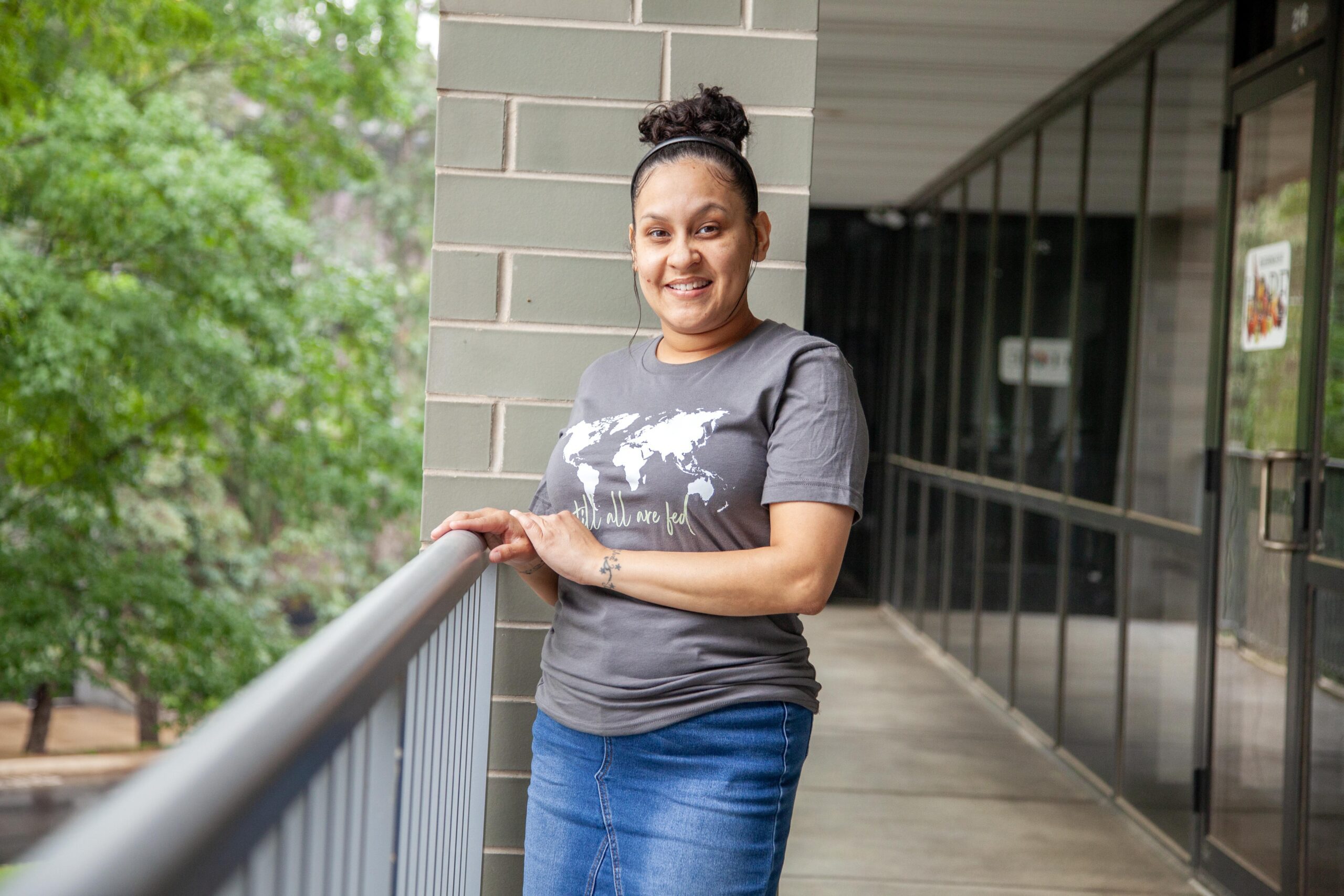 Woman smiling while leaning on a railing in a modern building with trees in the background, wearing a gray t-shirt featuring a world map and the phrase "until all are fed."
