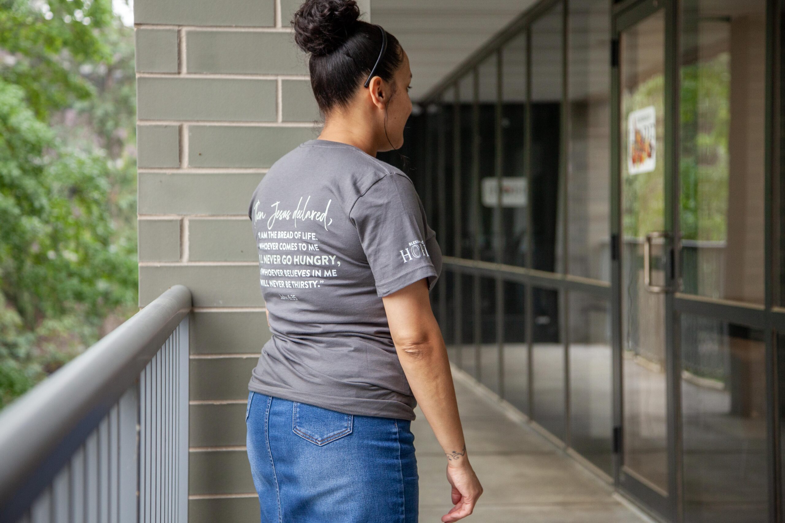 A woman stands on a balcony wearing a gray t-shirt with a religious message about Jesus, highlighting themes of nourishment and belief. The background features greenery and a building corridor.