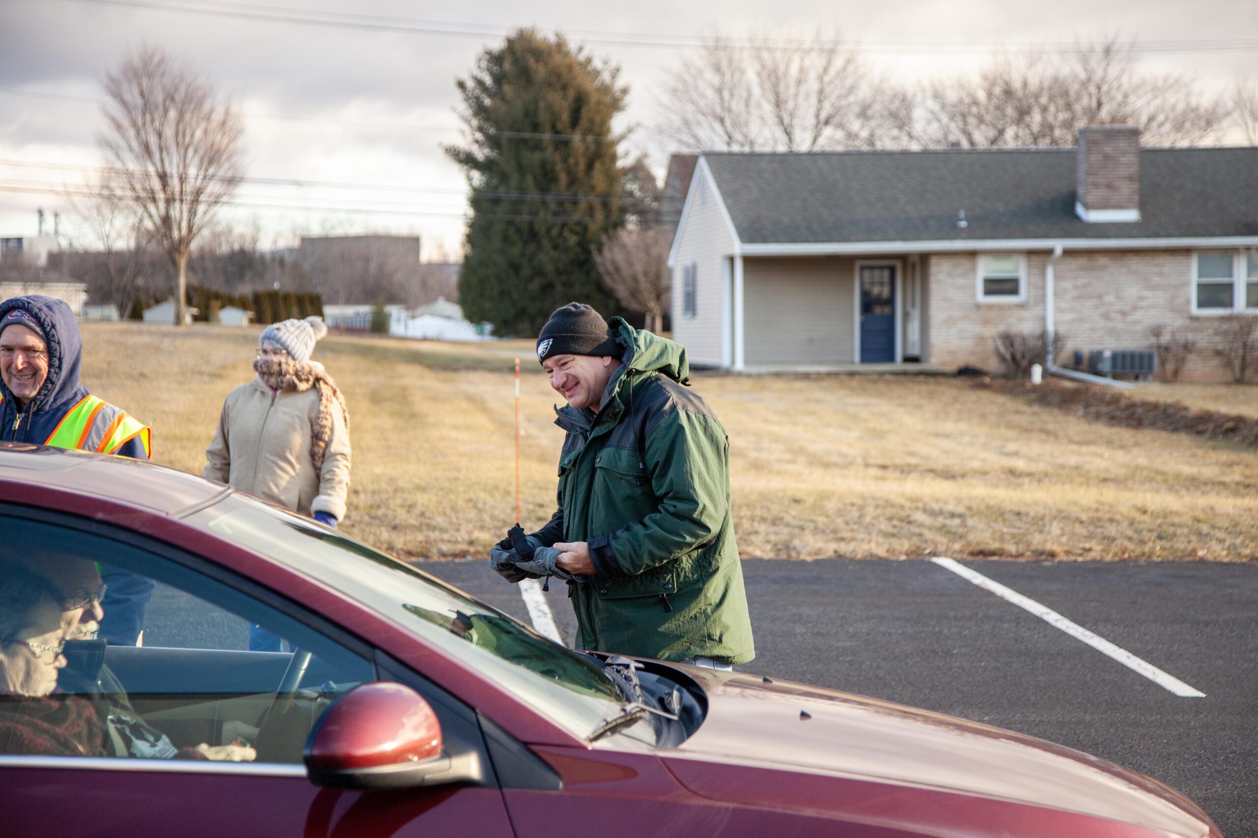 Image of a community event where volunteers are assisting individuals in a car. One man wearing a green jacket smiles as he approaches the car, while another volunteer stands nearby wearing a reflective vest. In the background, a residential area is visible with a house and trees, indicating a winter setting.