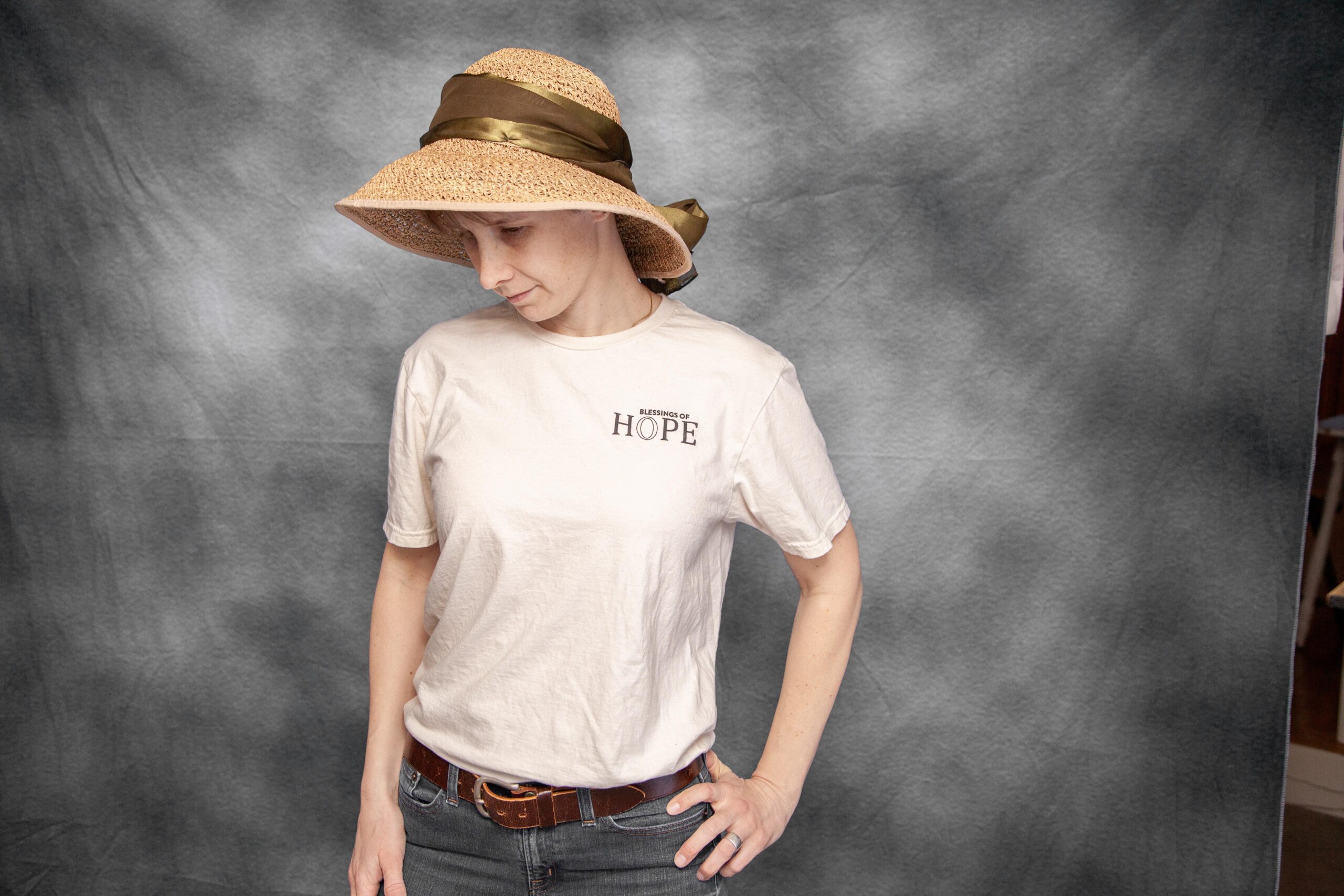 Woman wearing a beige t-shirt with "Blessings of Hope" design and a wide-brimmed straw hat with a brown ribbon, standing against a gray backdrop. The image conveys a sense of positivity and resilience.