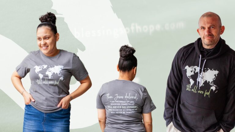 Two individuals wearing matching apparel with a world map graphic, promoting the message "Till all are fed." The woman is in a gray t-shirt showcasing a quote from the Bible, while the man wears a black hoodie with the same graphic. The background features a soft, abstract design, emphasizing their cause and mission with Blessings of Hope.