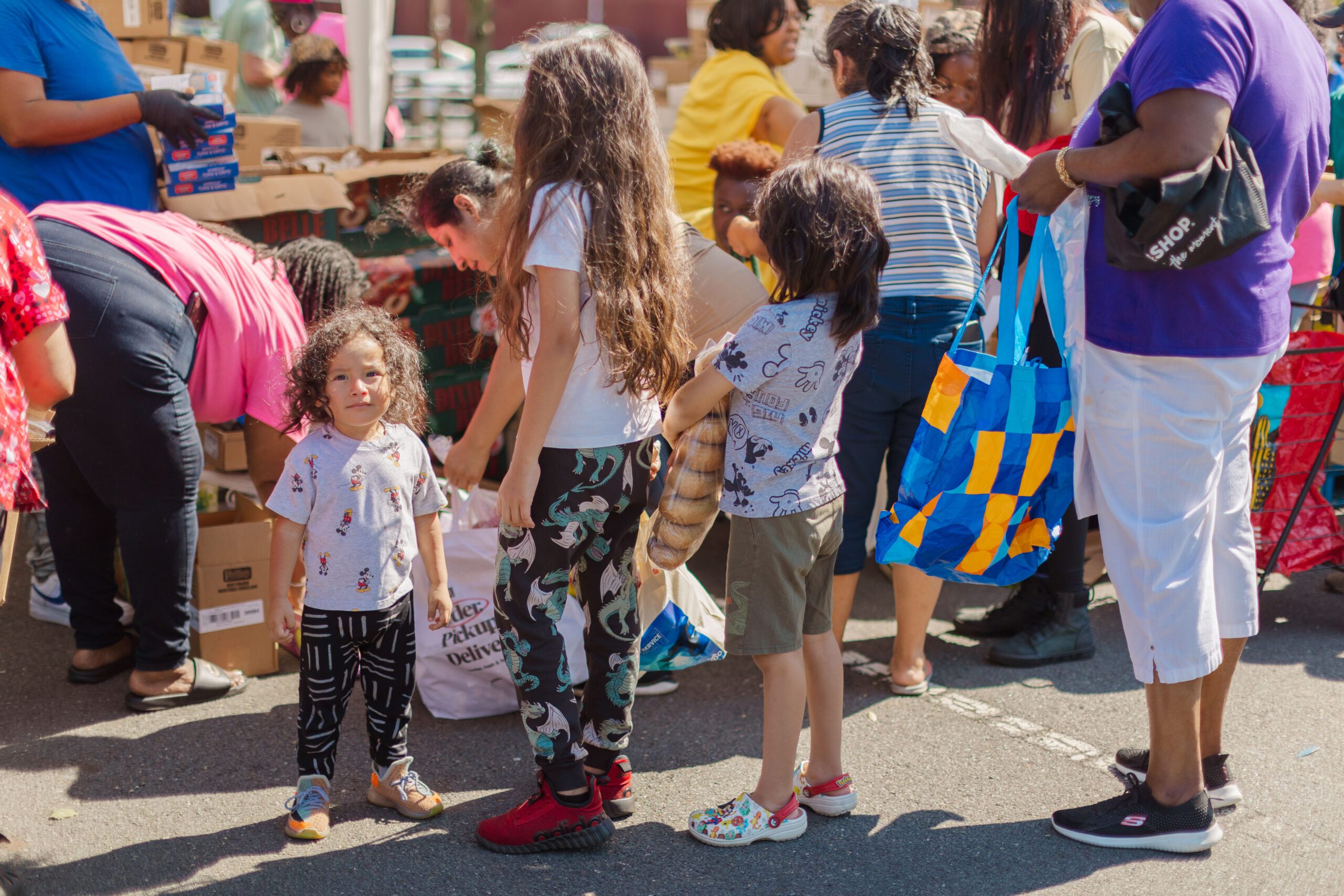 Children engaging in a community food distribution event, with adults organizing boxes and handing out food. The scene highlights diversity and support within the community, featuring kids in casual clothing and a festive atmosphere.