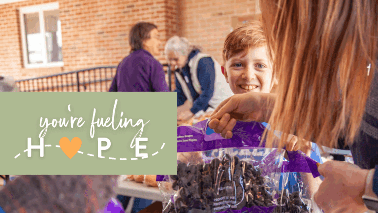 A young boy smiles while receiving a bag of seedless grapes from a volunteer during a community food distribution event. The backdrop features individuals engaging in conversation, emphasizing community support and helping to fuel hope through shared efforts.