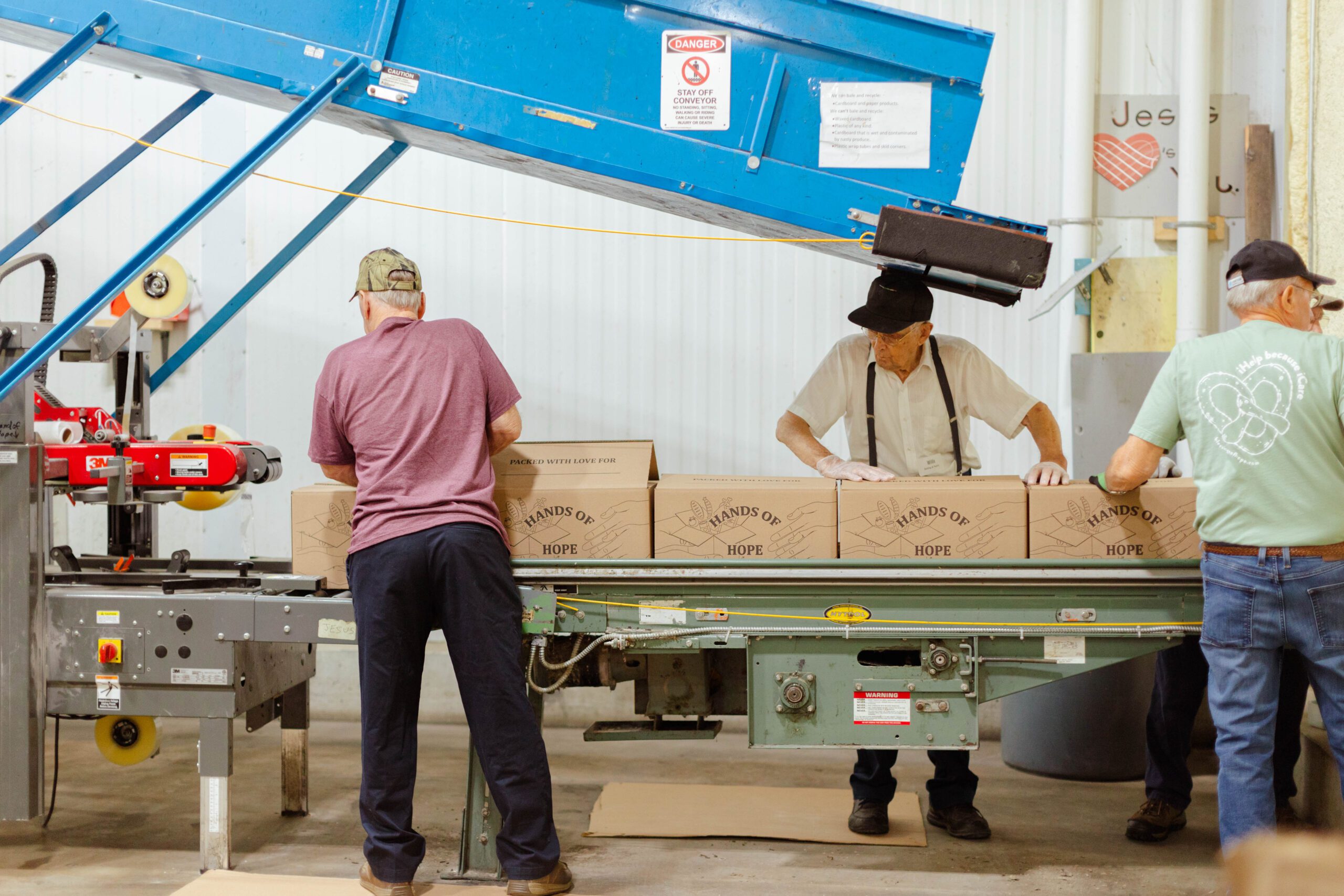 Volunteers packing boxes labeled "Hands of Hope" at a conveyor in a warehouse, demonstrating community support and teamwork in a charitable effort.
