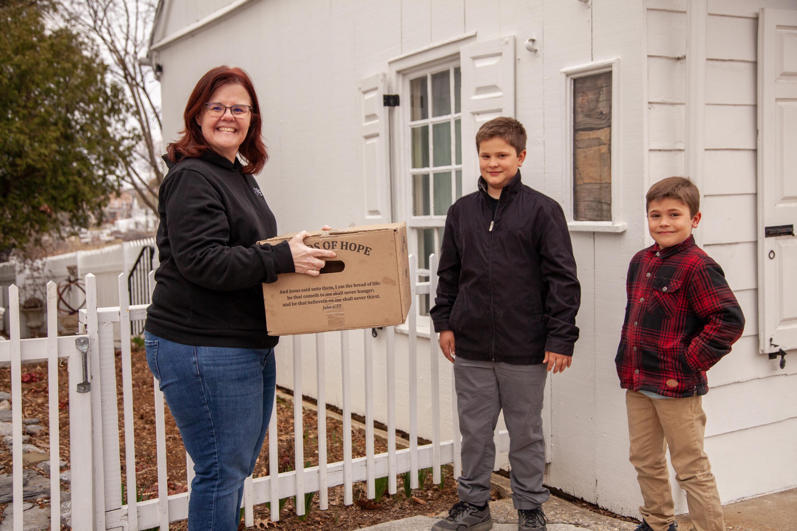 A woman and two boys stand outside a house, with the woman smiling and holding a box labeled "Boxes of Hope." The boys are dressed in jackets, one wearing a plaid shirt, and they are positioned beside her in a friendly demeanor. The setting features a white fence and a quaint home backdrop, symbolizing community support and generosity.