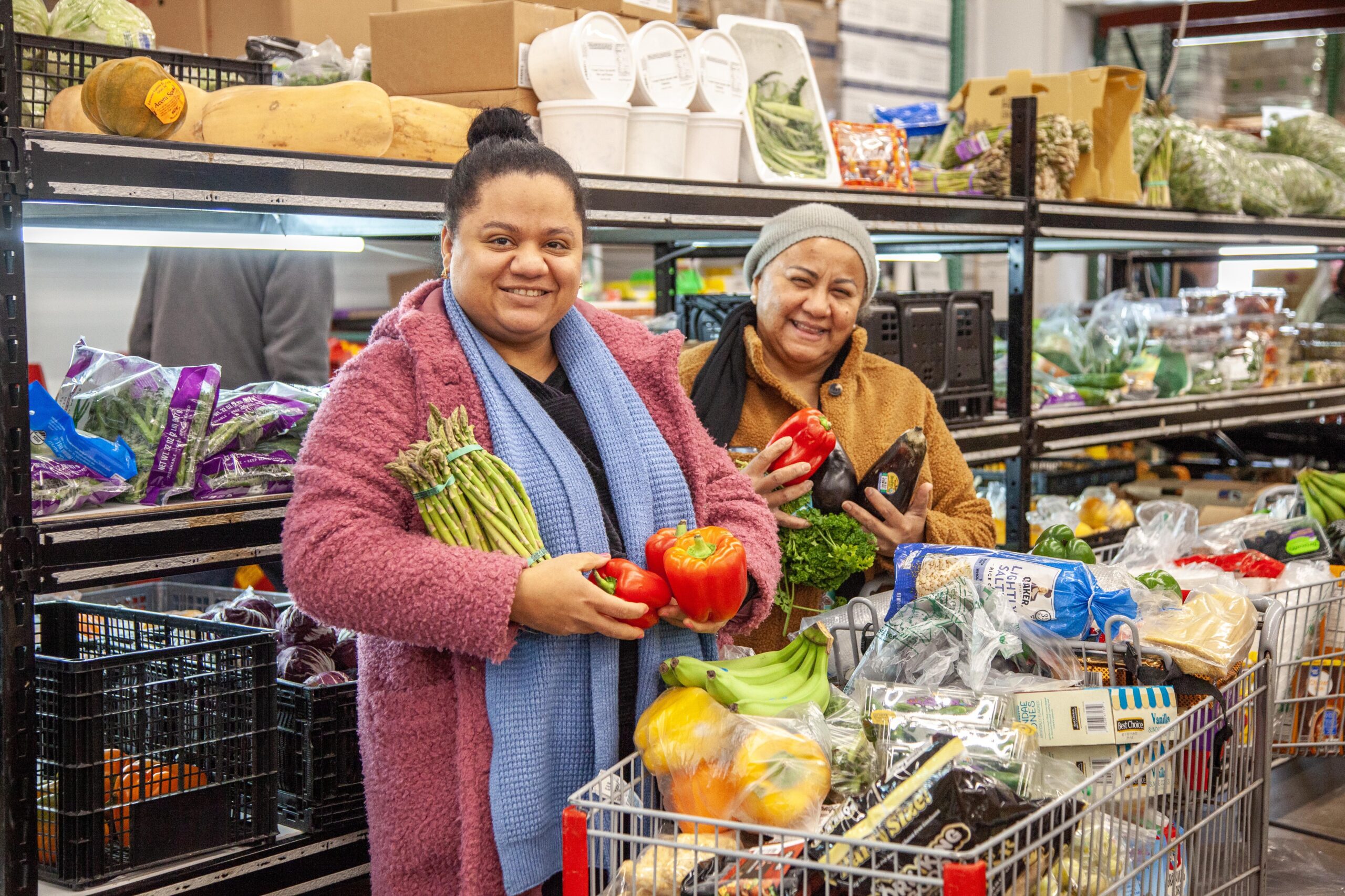 Two women shopping for fresh vegetables at a market, smiling as they hold vibrant red peppers and asparagus, surrounded by a variety of produce in carts and on shelves.