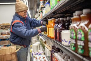 An employee wearing a winter hat and gloves restocks shelves with various beverage products in a grocery store aisle.
