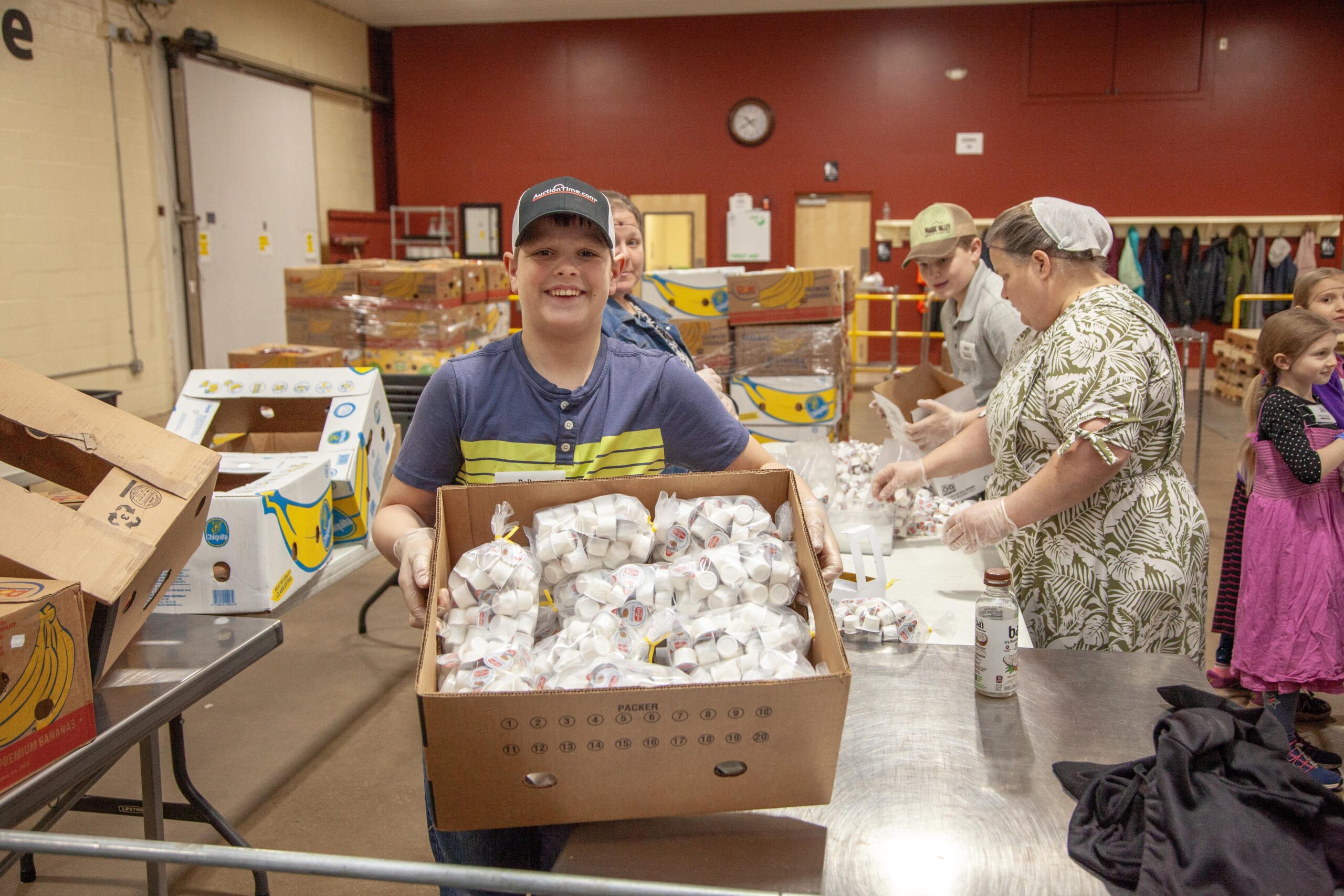 A group of volunteers participating in a food distribution event, with a young boy smiling and holding a box full of packaged food items. Other volunteers are seen in the background organizing supplies on tables in a community center setting.