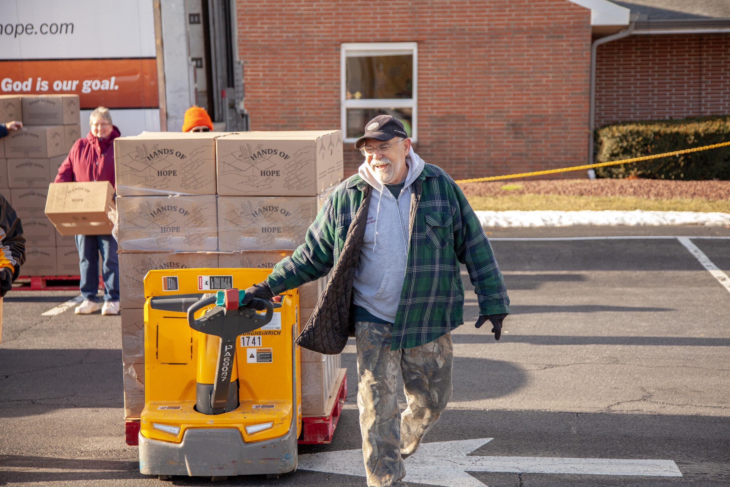 Man smiling while using a pallet jack to move boxes marked "Hands of Hope" at a community food distribution event, with volunteers assisting in the background.
