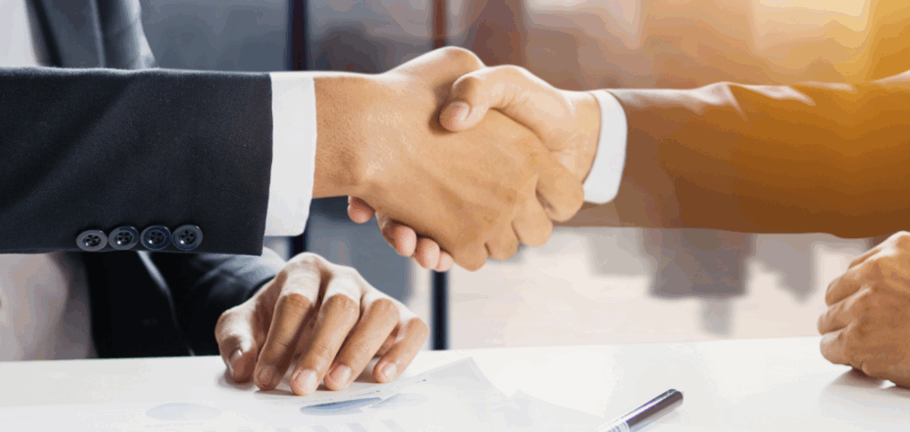 Business professionals shaking hands in a formal setting, symbolizing partnership and collaboration, with documents and a pen on the table.