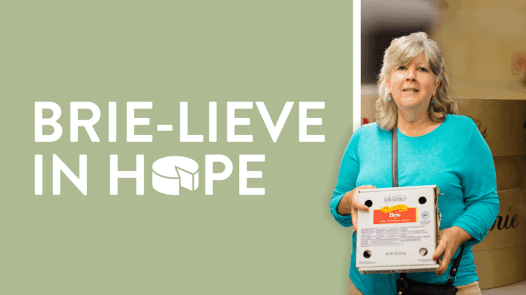 Woman holding a box of brie cheese with the text "Brie-lieve in Hope" in the background, promoting a charitable initiative related to food distribution.