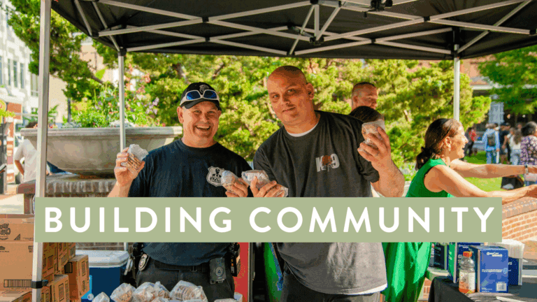 Two community members smiling and holding food packages at an outdoor event under a tent, promoting community engagement and support. In the background, others are interacting, surrounded by greenery and a vibrant atmosphere.
