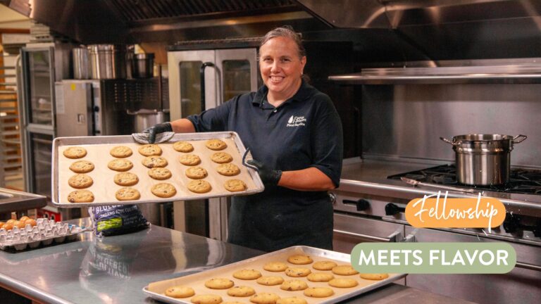 A smiling woman in a black shirt holds a tray of freshly baked chocolate chip cookies in a professional kitchen, showcasing the delicious treats on parchment paper. In the background, additional trays and kitchen equipment are visible, emphasizing a vibrant baking environment.