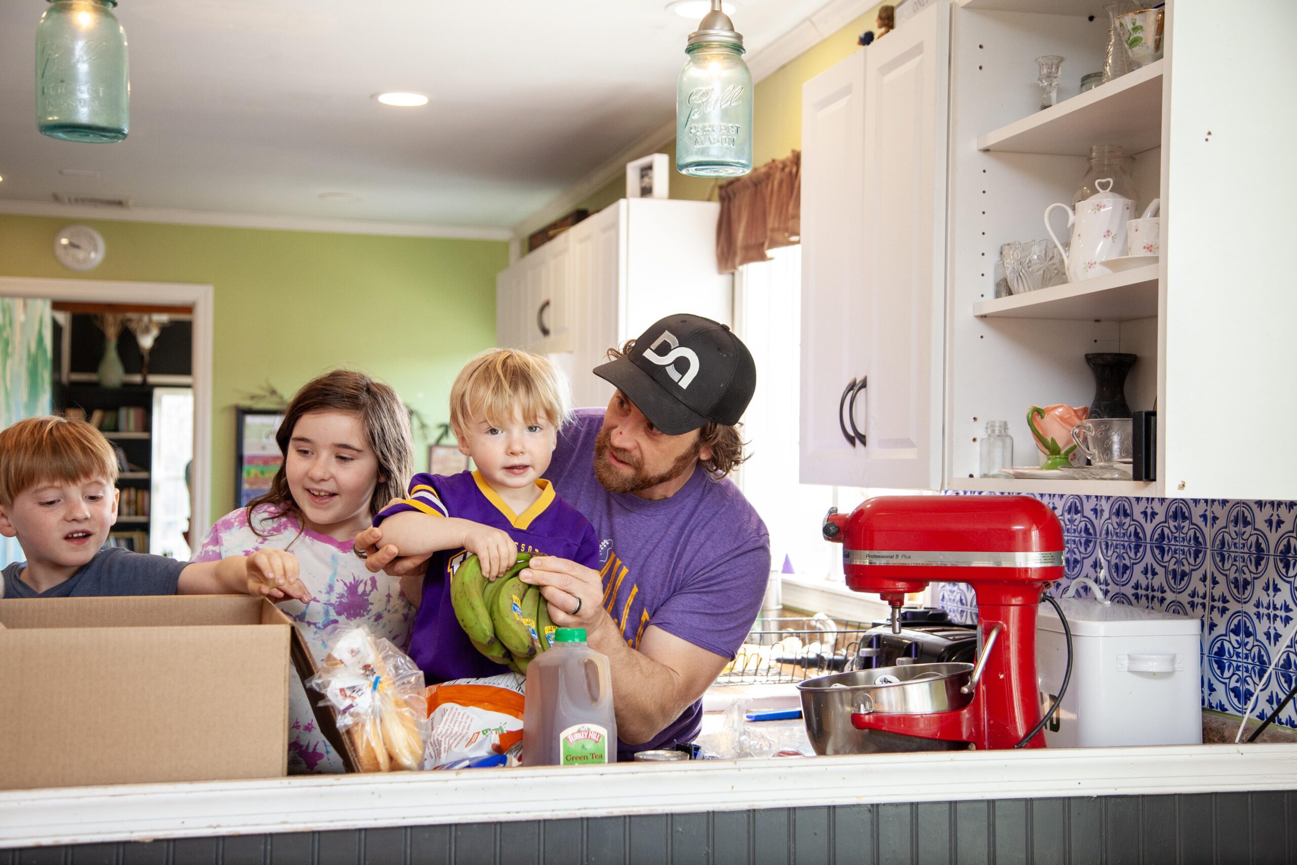 A father and three children engage in a fun cooking activity in a colorful kitchen, unpacking groceries from a cardboard box. The scene features a red stand mixer and various food items, highlighting a family bonding moment focused on cooking and togetherness.
