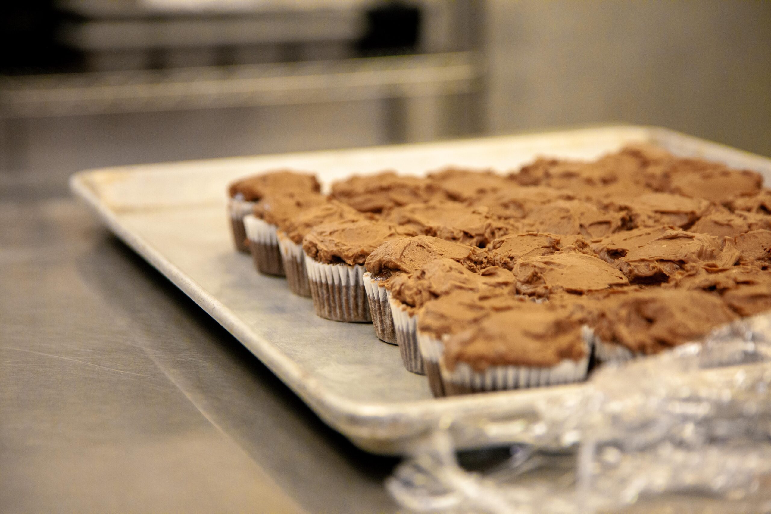 Freshly baked chocolate cupcakes with rich frosting arranged on a metal tray in a kitchen setting.