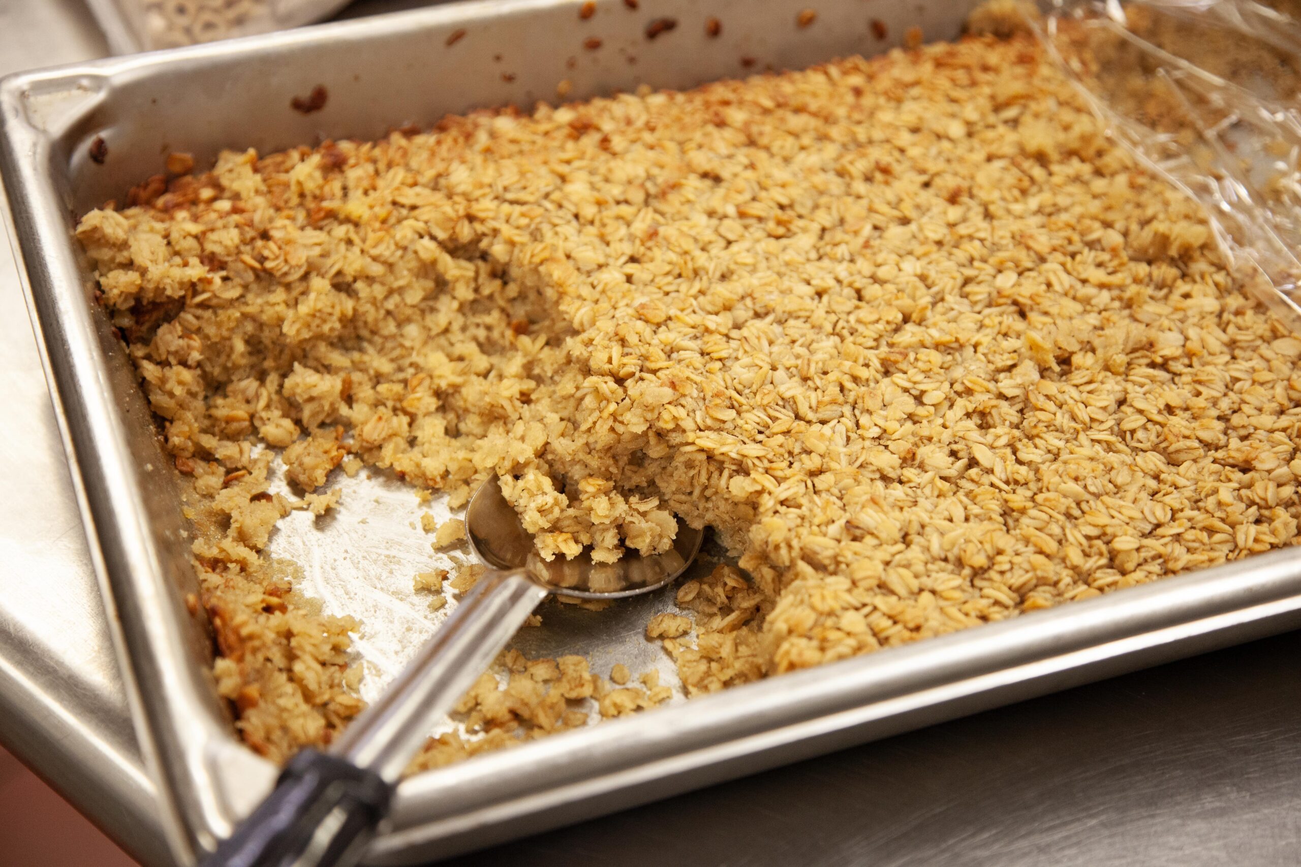 Baked oatmeal in a metal tray with a portion removed, showcasing a golden-brown top and a scoop resting beside it. Ideal for breakfast or healthy snacks.