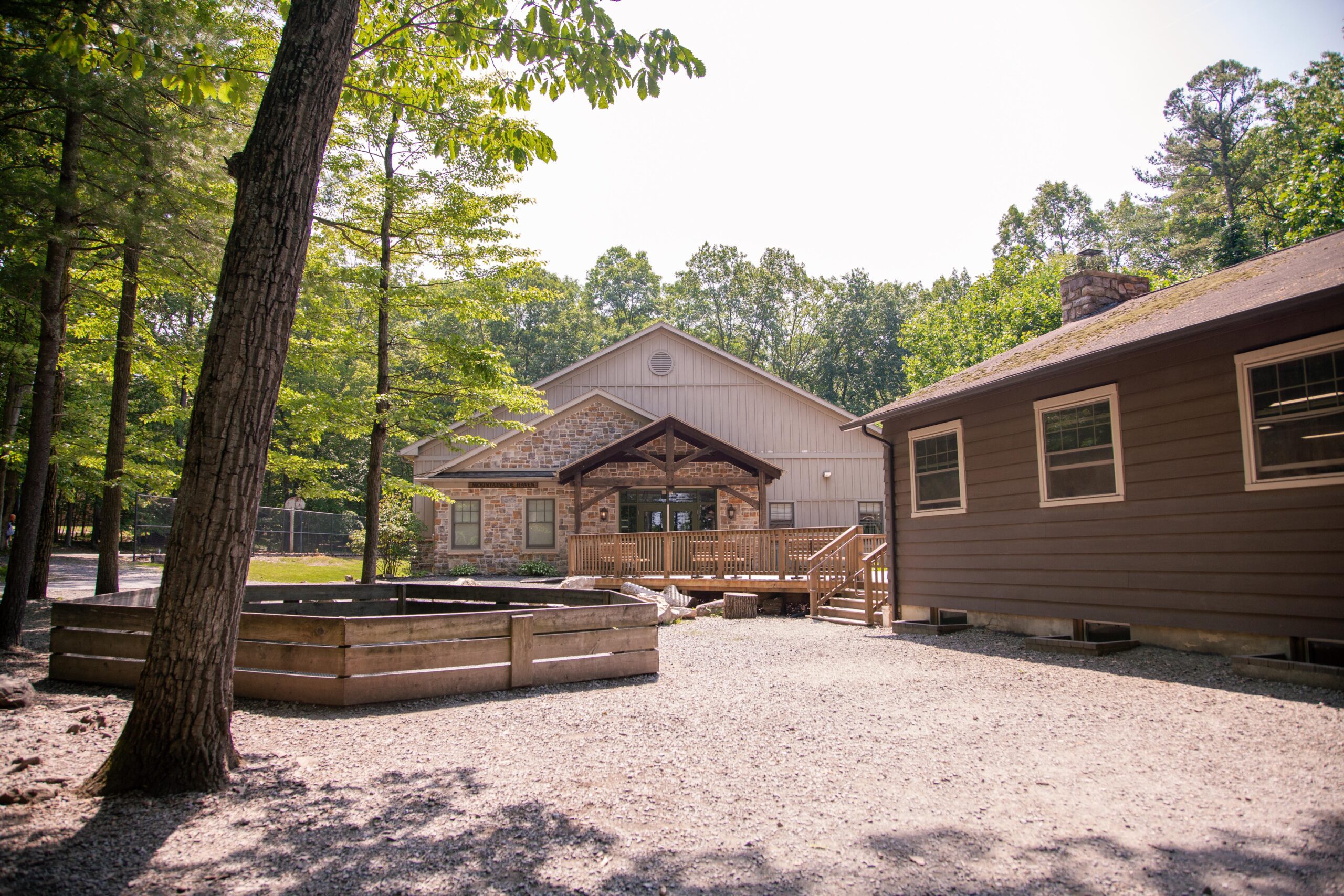 Exterior view of a rustic lodge surrounded by trees, featuring a wooden deck and gravel pathway, ideal for outdoor activities and gatherings.