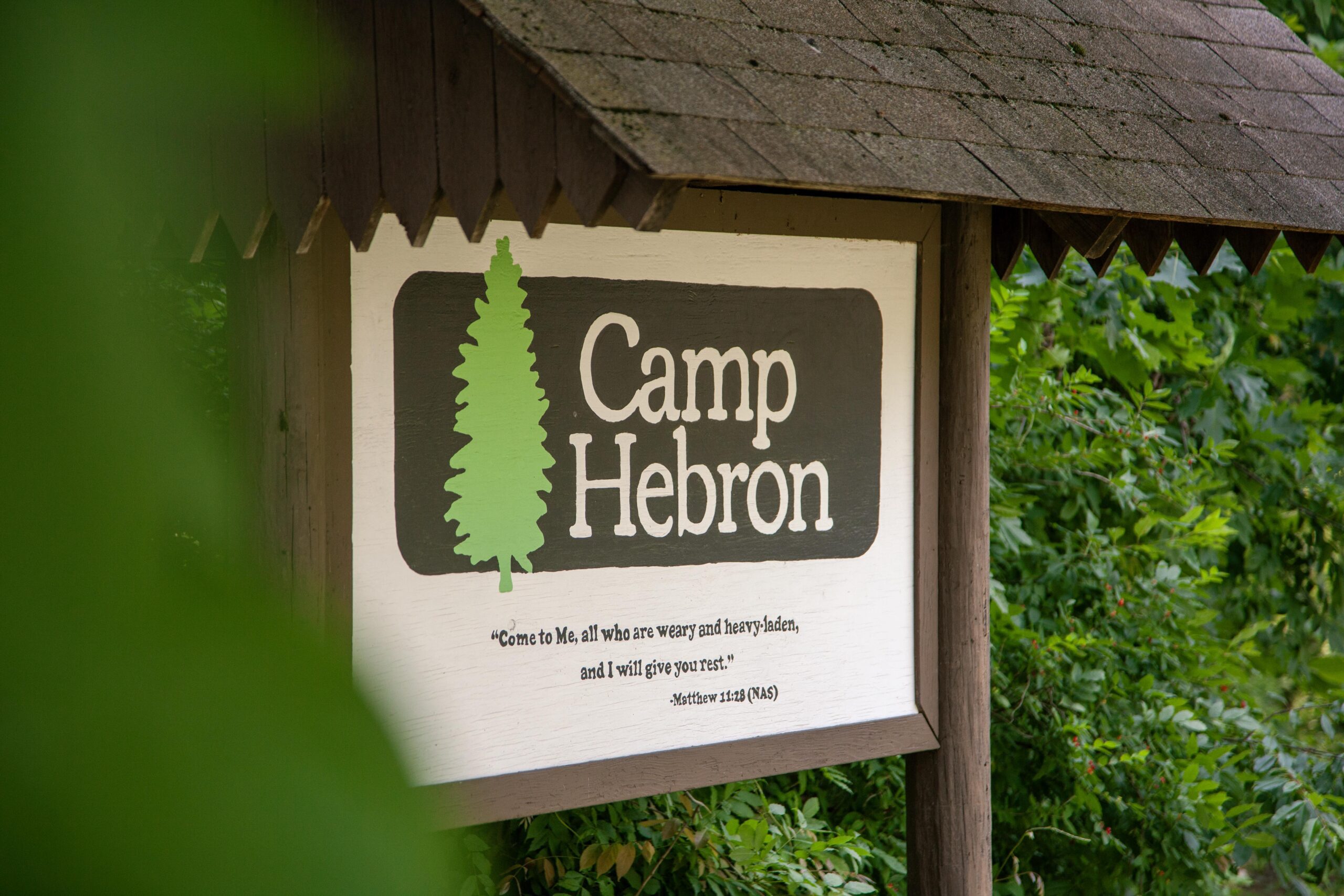 Sign for Camp Hebron featuring a green tree logo, surrounded by lush greenery, with a biblical quote encouraging rest and solace.