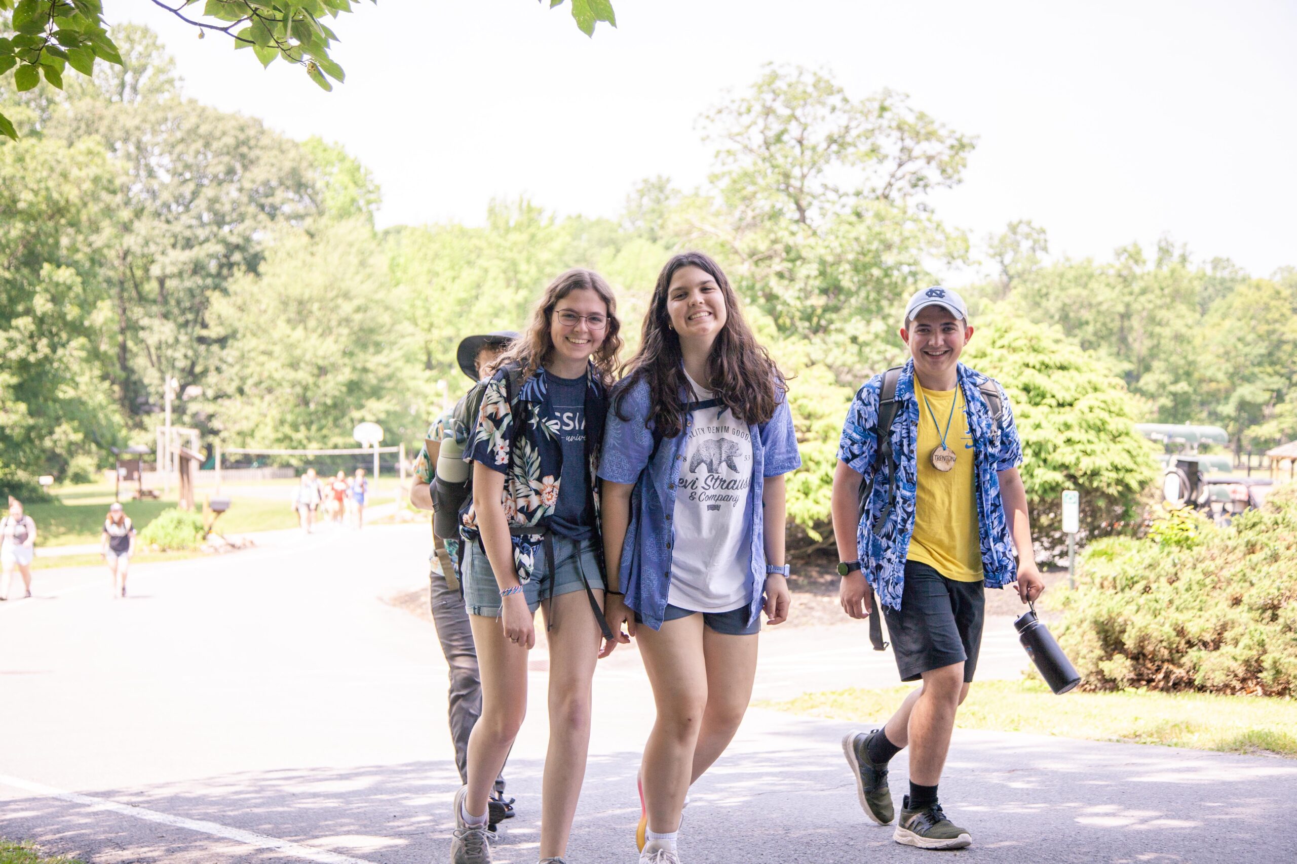 Group of young people walking along a sunny path in a park, wearing casual summer clothing, enjoying outdoor activities, and smiling. Lush green trees and other park visitors are visible in the background.