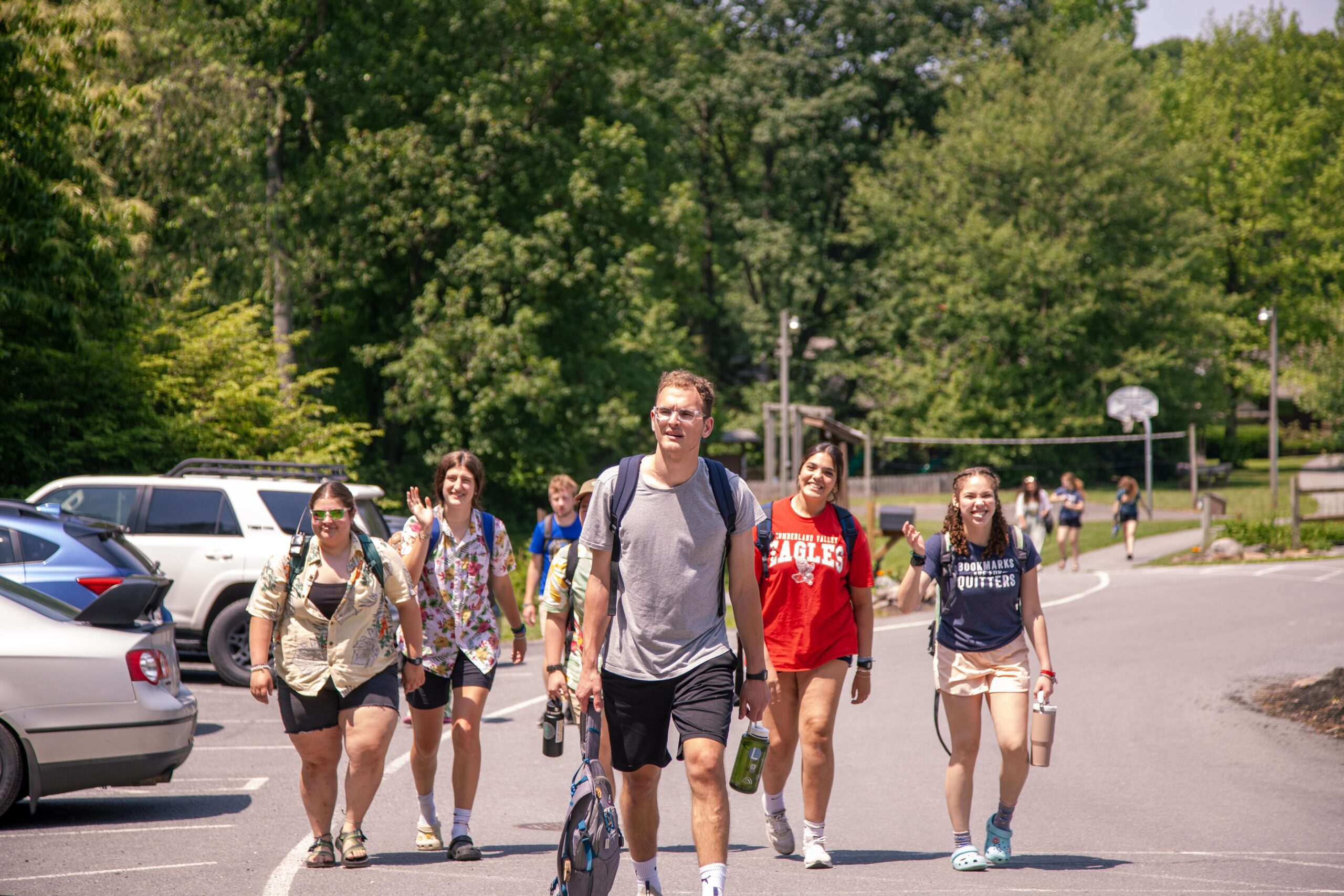 Group of six young adults walking together on a sunny day, carrying backpacks and water bottles, near a parking area surrounded by trees and recreational facilities.