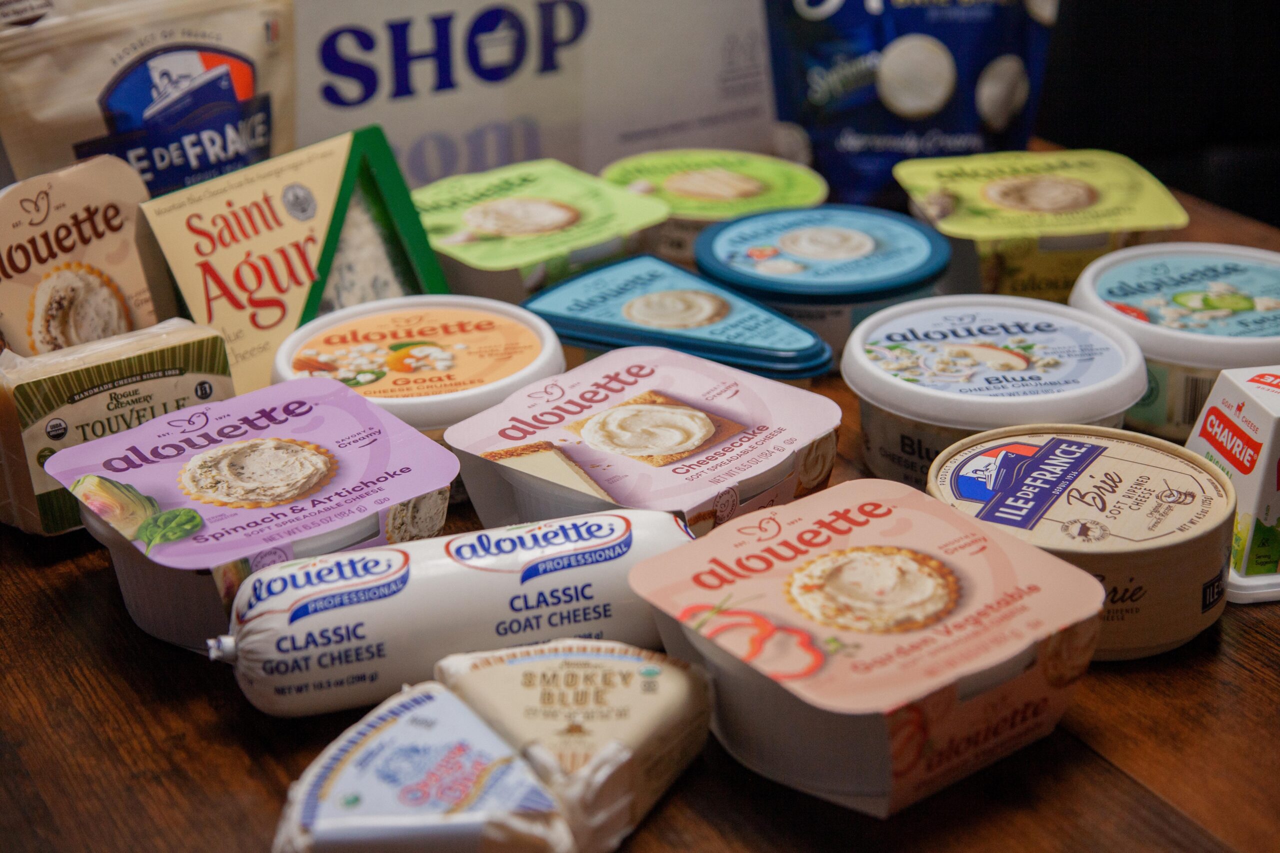 An assortment of various cheese products, including Alouette spreads in flavors like Spinach & Artichoke and Cheesecake, along with Ile de France Brie and Saint Agur cheese, displayed on a wooden surface.