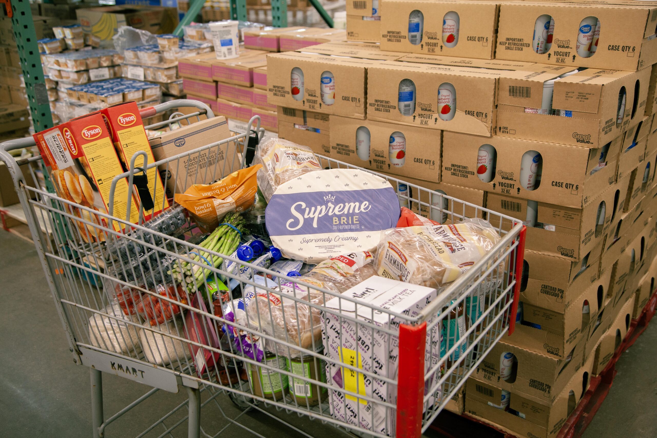 Shopping cart filled with a variety of grocery items, including Tyson chicken products, fresh vegetables like asparagus, and Supreme Brie cheese, set against stacks of packaged yogurt and other food items in a warehouse store.