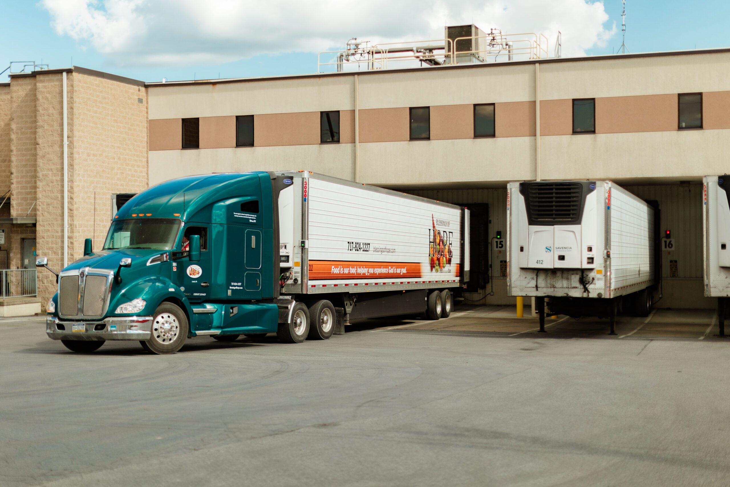 A teal semi-truck with a refrigerated trailer is parked at a loading dock next to two other refrigerated trailers. The backdrop features a commercial building with multiple loading bays, showcasing a busy logistics operation for food distribution.