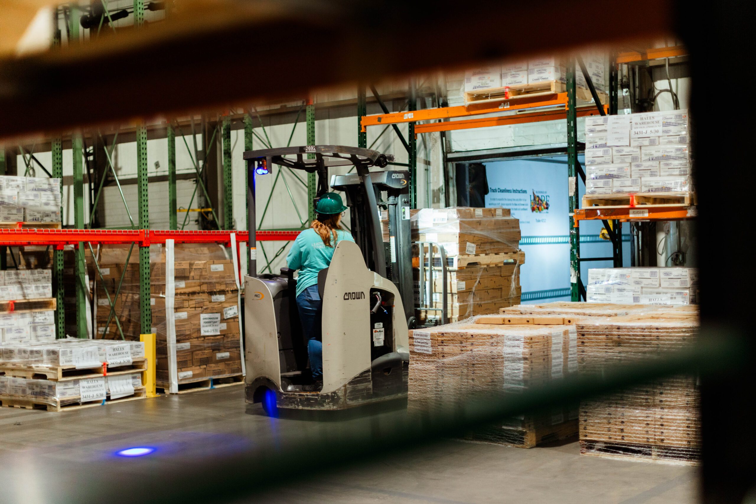 Warehouse worker operating a Crown forklift in a storage facility, surrounded by stacked pallets and organized shelving, highlighting efficient logistics and inventory management.