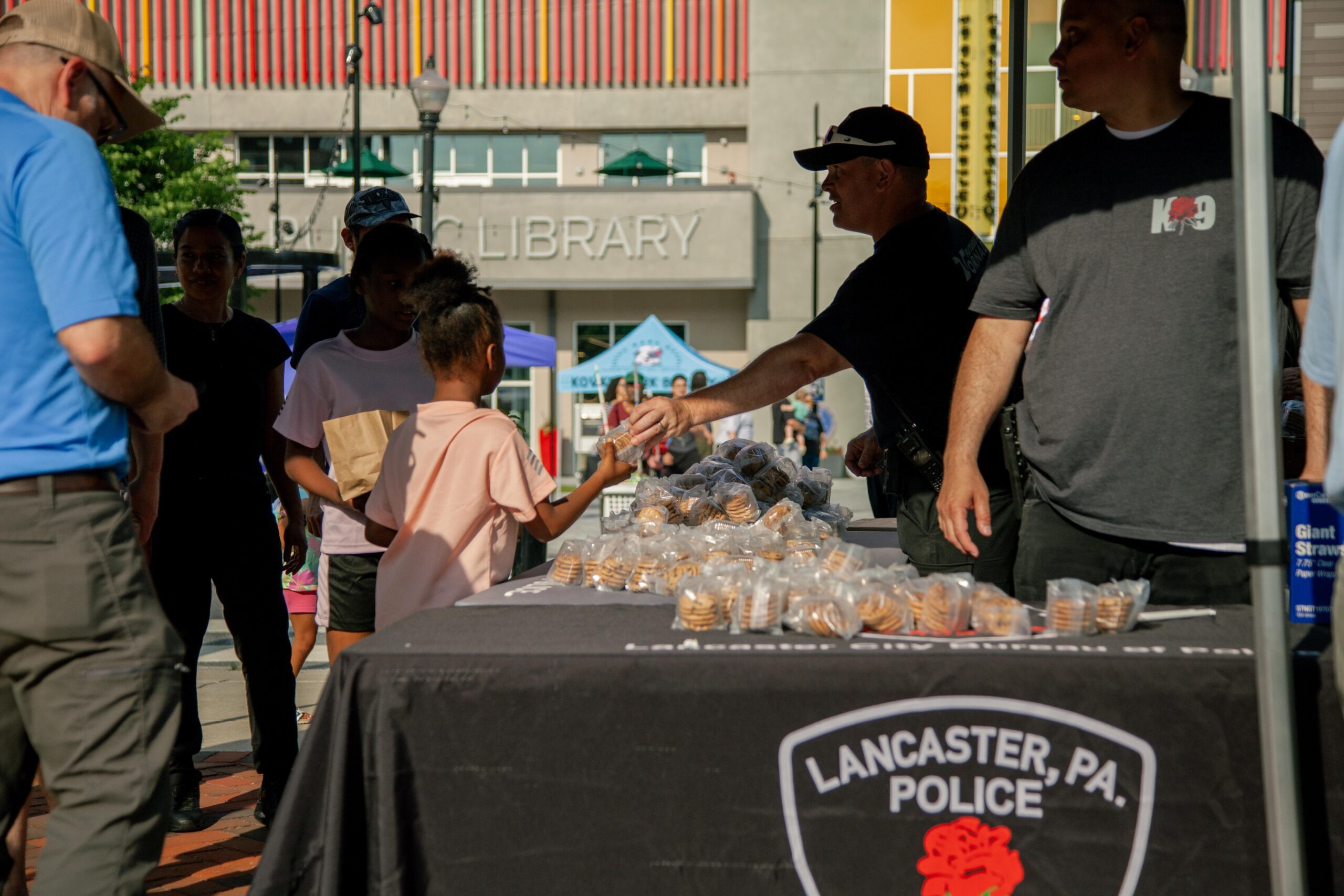 A police officer hands a cookie to a young child at a community event in front of the Lancaster Public Library, with a table displaying cookies and a banner featuring the Lancaster, PA Police Department.