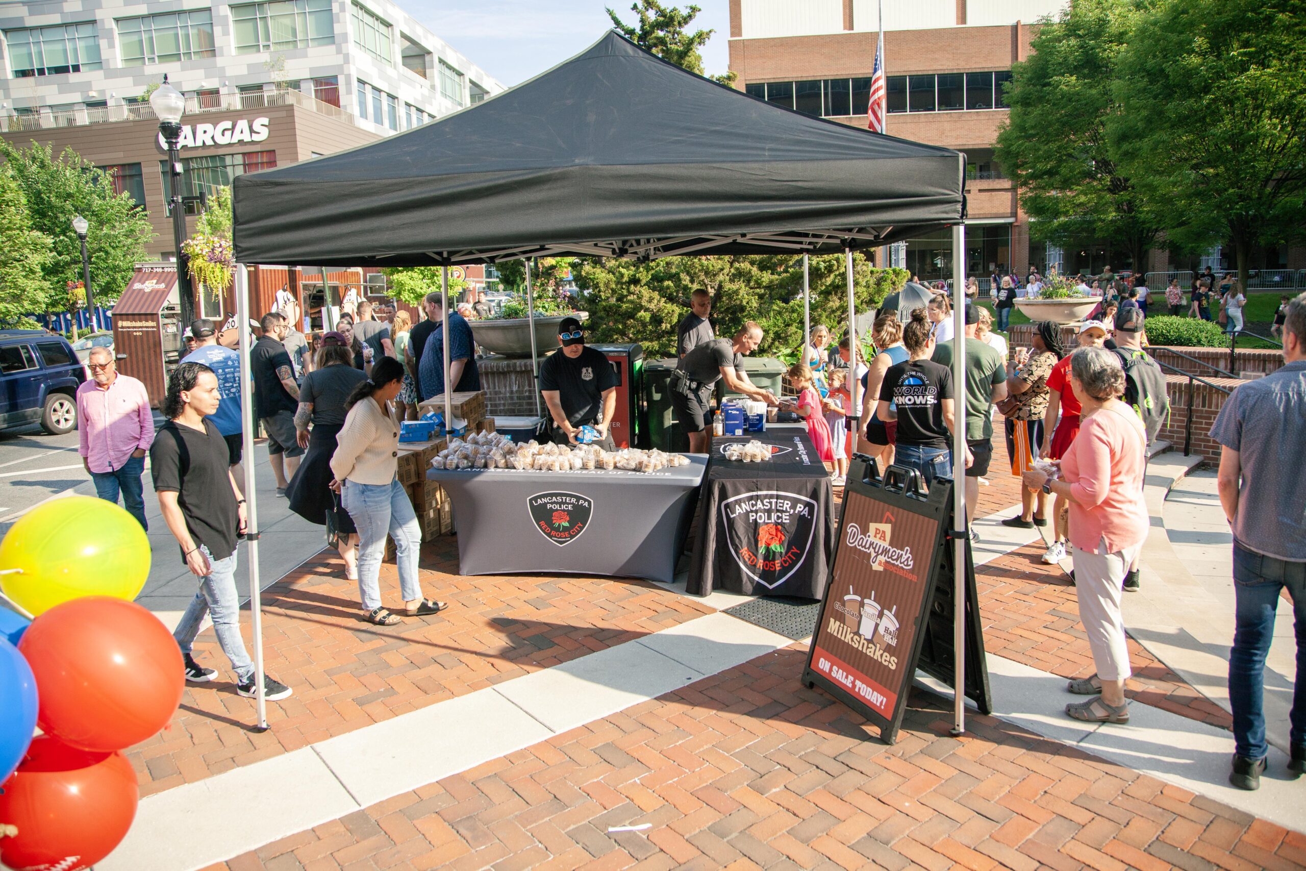 Crowd gathered at a community event featuring a tent with the Lancaster Police Department logo, offering food and drinks. Attendees enjoy the festivities, with colorful balloons in the foreground and a sign promoting milkshakes for sale.